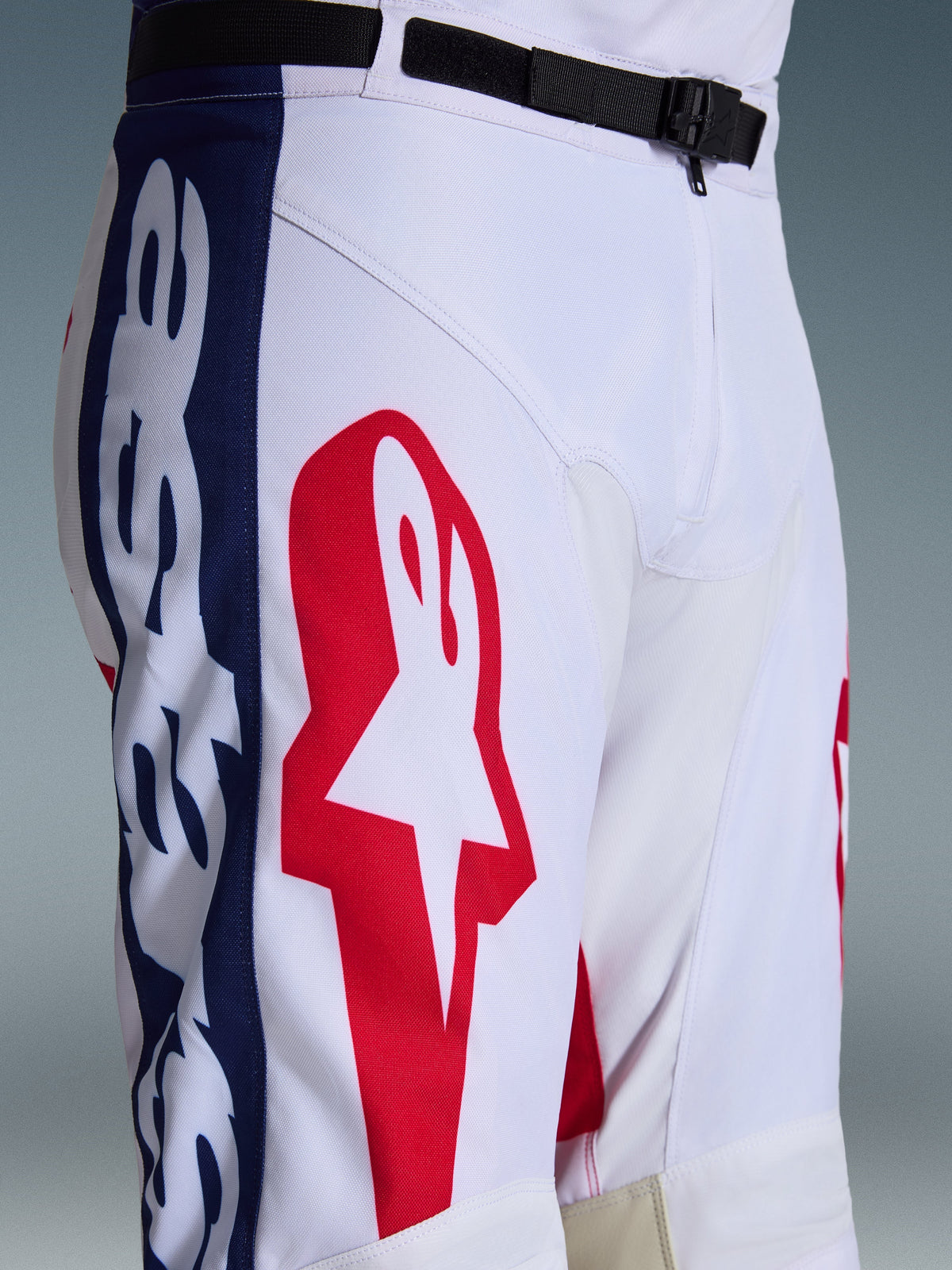 Alpinestars Racer Riway Pants 2026, spodnie motocrossowe, biało-niebiesko-czerwone, zbliżenie na górną część nogawki i talię pokazujące duże czerwone logo gwiazdy na białym materiale poliestrowym, niebieskie panele boczne z brandingiem oraz czarny regulowany pasek w pasie z magnetyczną klamrą SX2, sportowy design wyścigowy off-road