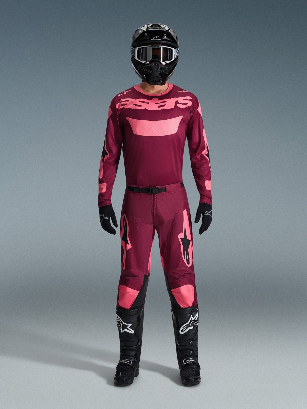 Motocross rider wearing complete racing gear, Dark Red Coral jersey with pink accents, matching Racer Riway pants, black helmet with white details, black boots with white logos, black gloves, coordinated outfit for off-road racing