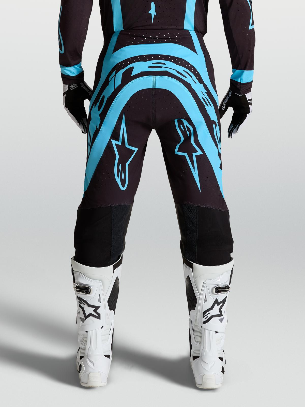 Alpinestars Spodnie Supertech Vesta, spodnie motocrossowe off-road, czarne i turkusowe, widok z tyłu z białymi butami jeździeckimi, profesjonalna odzież wyścigowa