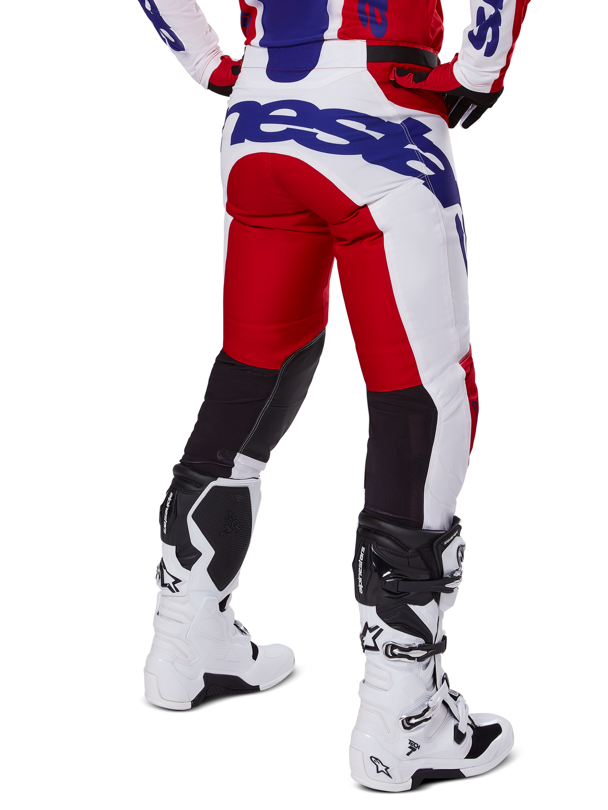 Alpinestars 2025 Racer Veil Pants, spodnie motocrossowe, jasnoczerwone i białe z niebieskim brandingiem, widok z tyłu ukazujący czarne elastyczne panele wentylacyjne z tyłu nogawek, zestawione z białymi butami motocrossowymi.