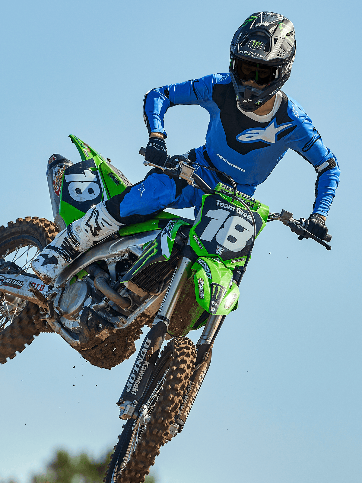Motocross rider mid-jump on a green motorcycle, wearing Alpinestars 2025 Racer Veil Pants in Blue and Black, matching blue jersey with white chest logo, white and black boots, and black helmet, high-performance off-road gear for racing.