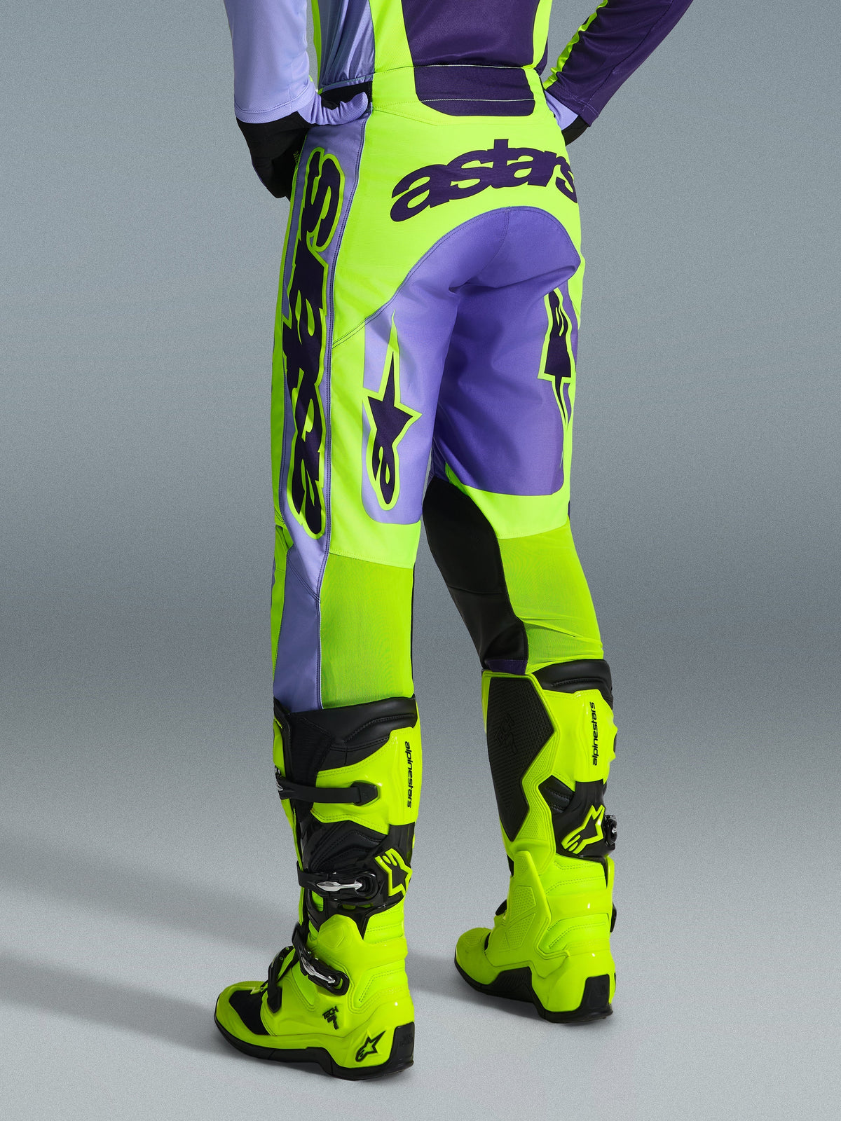 Alpinestars 2026 Racer Portl Pants, spodnie motocrossowe, fioletowe i żółte neonowe, widok z tyłu z wyraźnym brandingiem astars i fioletowymi akcentami, noszone z dopasowanymi żółtymi neonowymi butami motocrossowymi, wytrzymały materiał syntetyczny do wyścigów off-road
