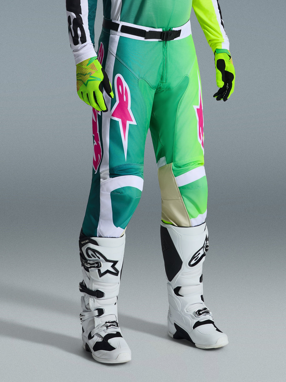 Alpinestars 2026 Racer Portl Pants, spodnie motocrossowe, zielone, białe i czarne z różowymi logo, noszone z białymi butami i neonowo-żółtymi rękawicami, wyraziste wzory graficzne, regulowana czarna klamra w pasie, odzież wyczynowa do jazdy w terenie