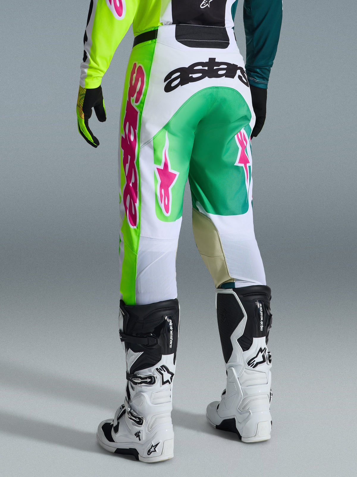 Spodnie Alpinestars Racer Portl 2026, spodnie motocrossowe, zielone, białe i czarne z różowymi akcentami, widok z tyłu z wyraźnym logo „astars” na siedzisku i różowymi logo gwiazdy na nogawkach, w połączeniu z biało-czarnymi butami motocrossowymi, profesjonalna odzież do wyścigów off-road