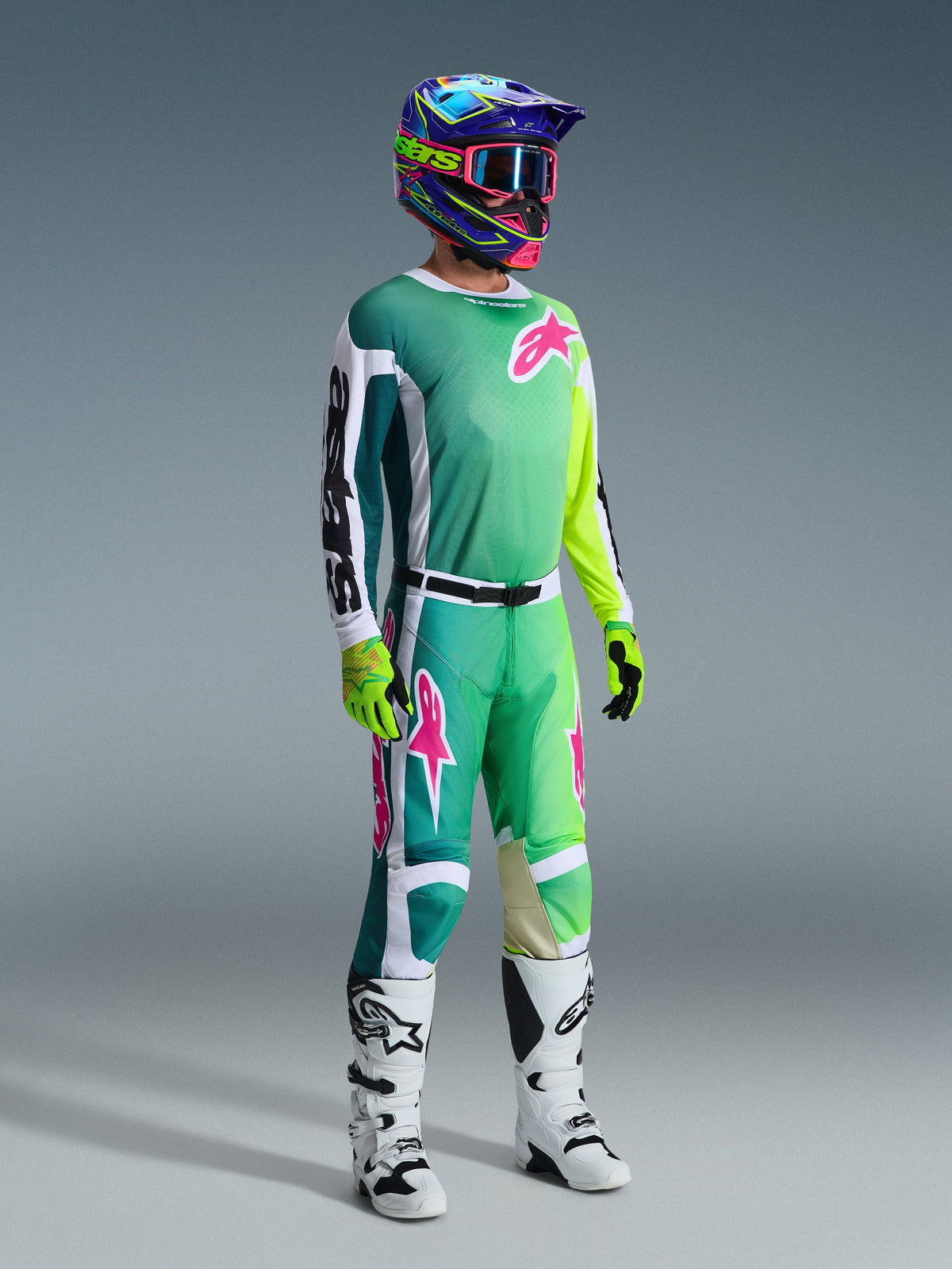 Motocyklista motocrossowy ubrany w spodnie Alpinestars 2026 Racer Portl i dopasowaną koszulkę w kolorach zielonym, białym i czarnym z różowymi akcentami logo, kolorowy kask, biało-czarne buty wyścigowe i neonowo-zielone rękawice, kompletny zestaw odzieży do jazdy off-road
