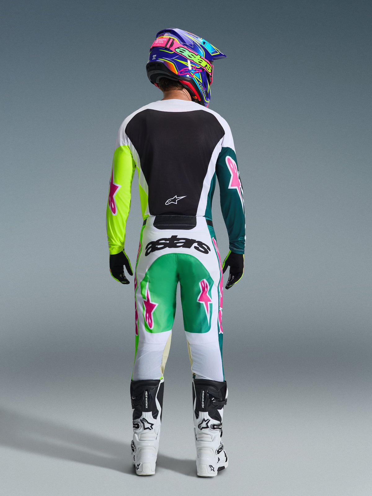 Zawodnik motocrossowy widziany od tyłu w spodniach Alpinestars 2026 Racer Portl w kolorze zielono-biało-czarnym, pasująca koszulka z różowymi logo gwiazdy, wielokolorowy kask 'astars' i biało-czarne buty, kompletny zestaw odzieży wyścigowej off-road.