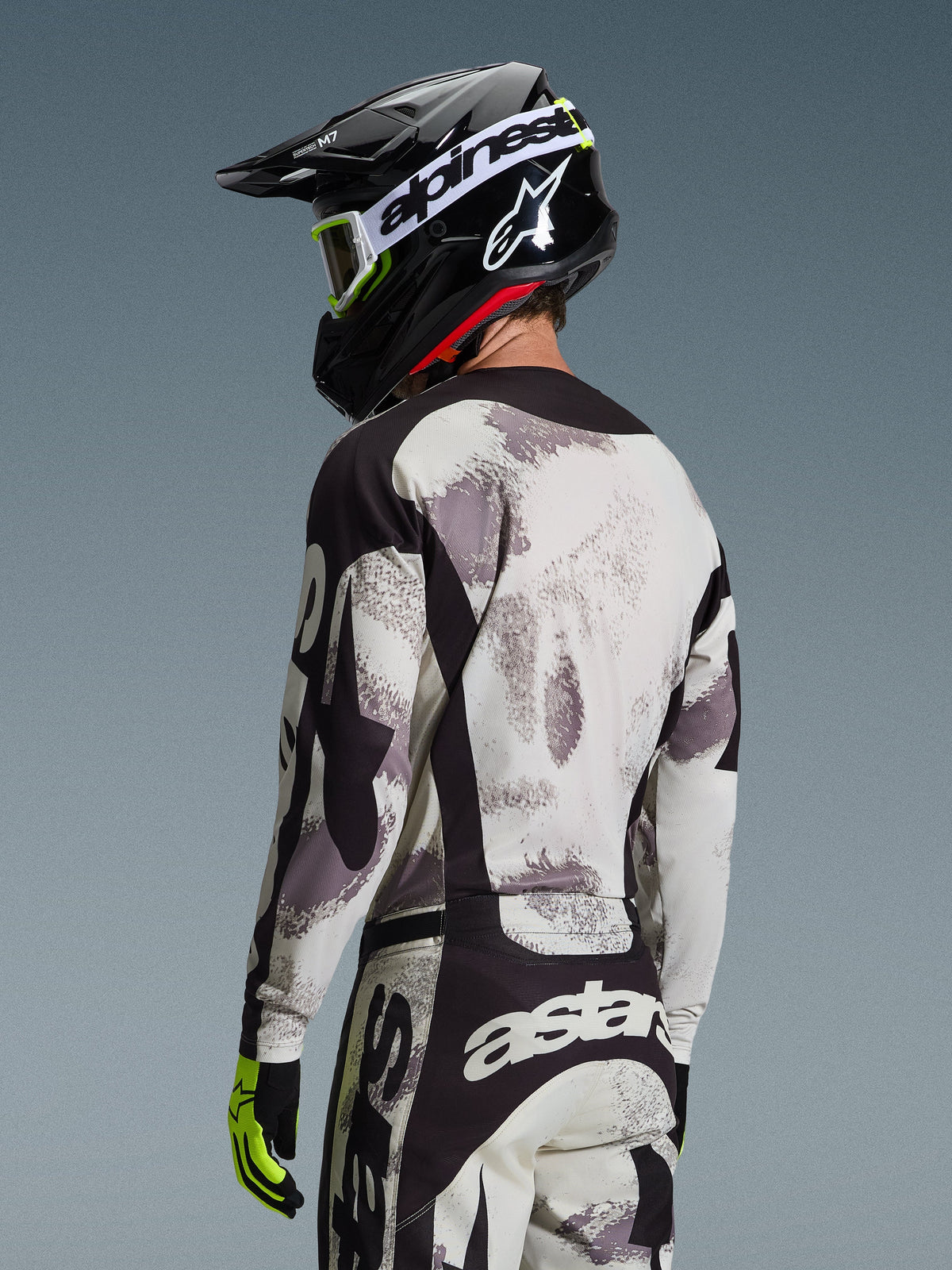 Zawodnik motocrossowy od tyłu w zestawie Alpinestars 2026 Racer Lahnd, koszulka i spodnie w szaro-białym kamuflażu Sand Camo z czarnymi akcentami, czarny kask z białym logo Alpinestars, neonowożółte i czarne rękawice, profesjonalny strój off-road