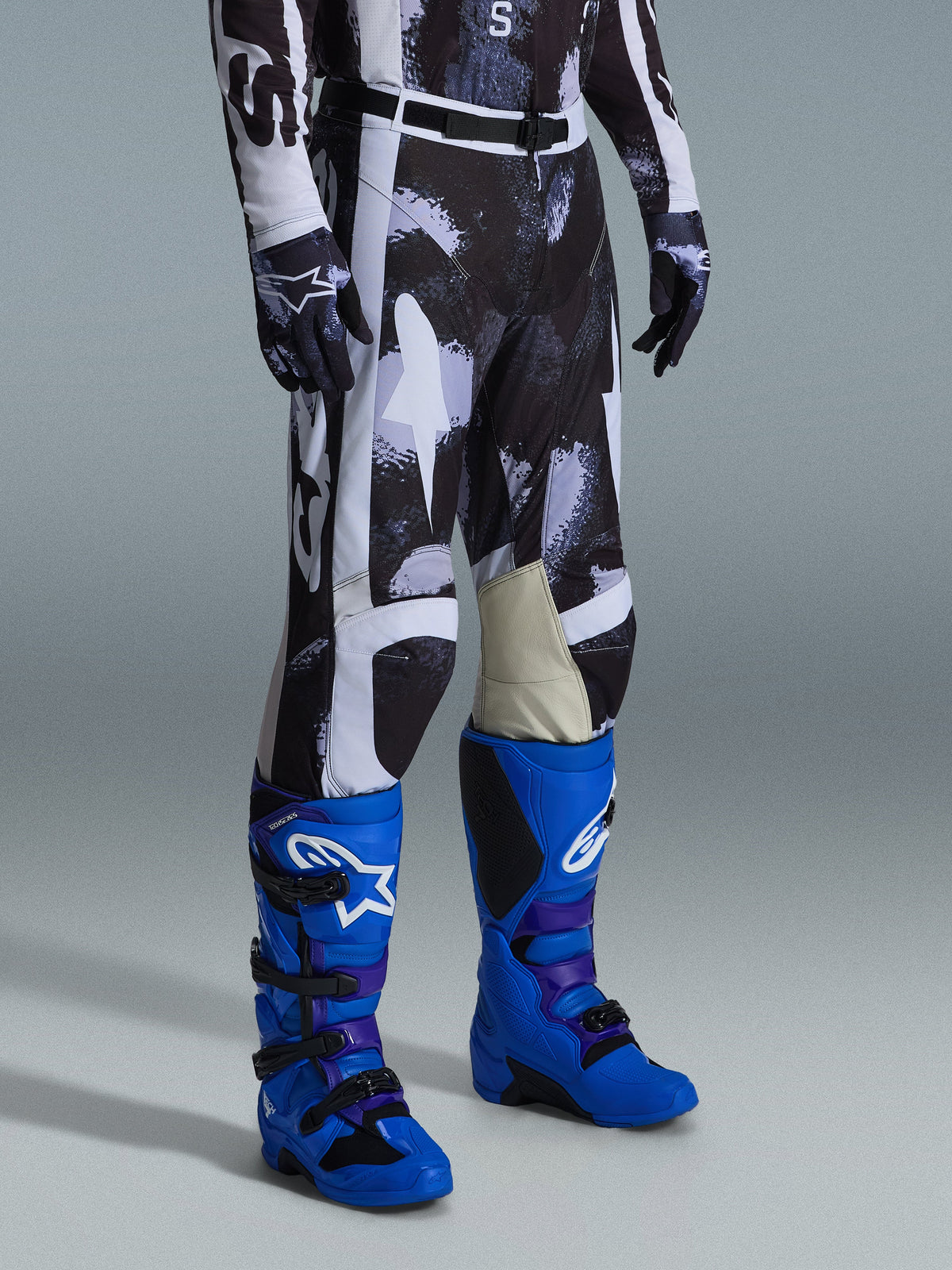 Alpinestars 2026 Racer Lahnd Pants, spodnie motocrossowe, szaro-czarny wzór Iron Camo, białe panele i wewnętrzna część kolan ze skóry koziej, zestawione z jaskrawoniebieskimi butami wyścigowymi i dopasowanymi rękawicami, odzież off-road o wysokiej wydajności