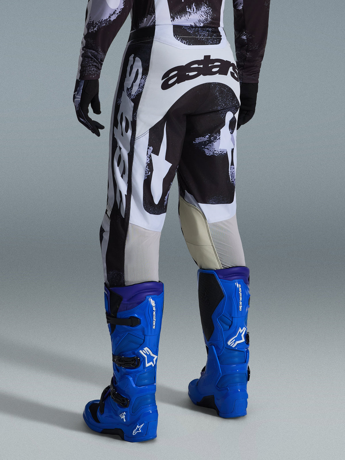 Alpinestars 2026 Racer Lahnd Pants, spodnie motocrossowe, szaro-czarny wzór Iron Camo, widok z tyłu z dużym biało-czarnym logo astars, w połączeniu z jaskrawoniebieskimi butami motocrossowymi, profesjonalny sprzęt do wyścigów off-road