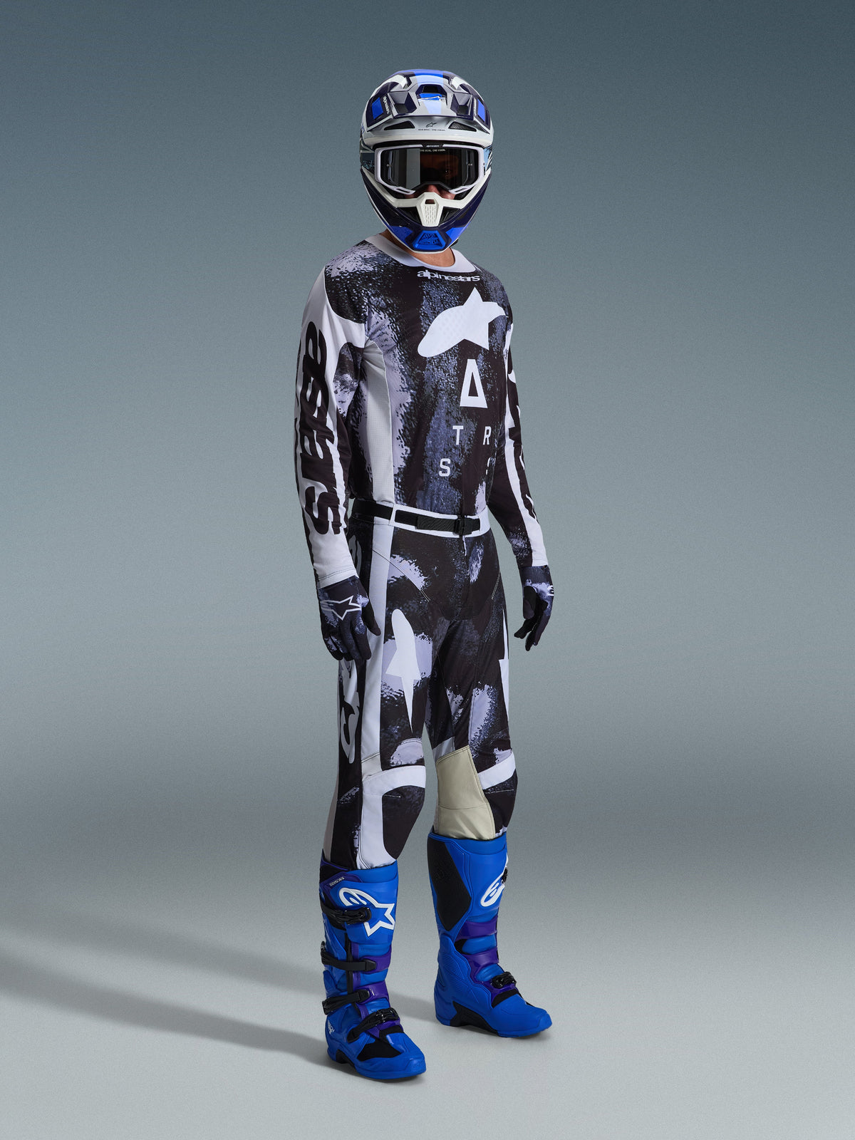 Motocyklista motocrossowy w pełnym rynsztunku, spodnie Alpinestars 2026 Racer Lahnd we wzorze Iron Camo i szarym, pasująca koszulka, jasnoniebieskie i fioletowe buty motocrossowe, niebiesko-biały kask, profesjonalny strój do wyścigów terenowych