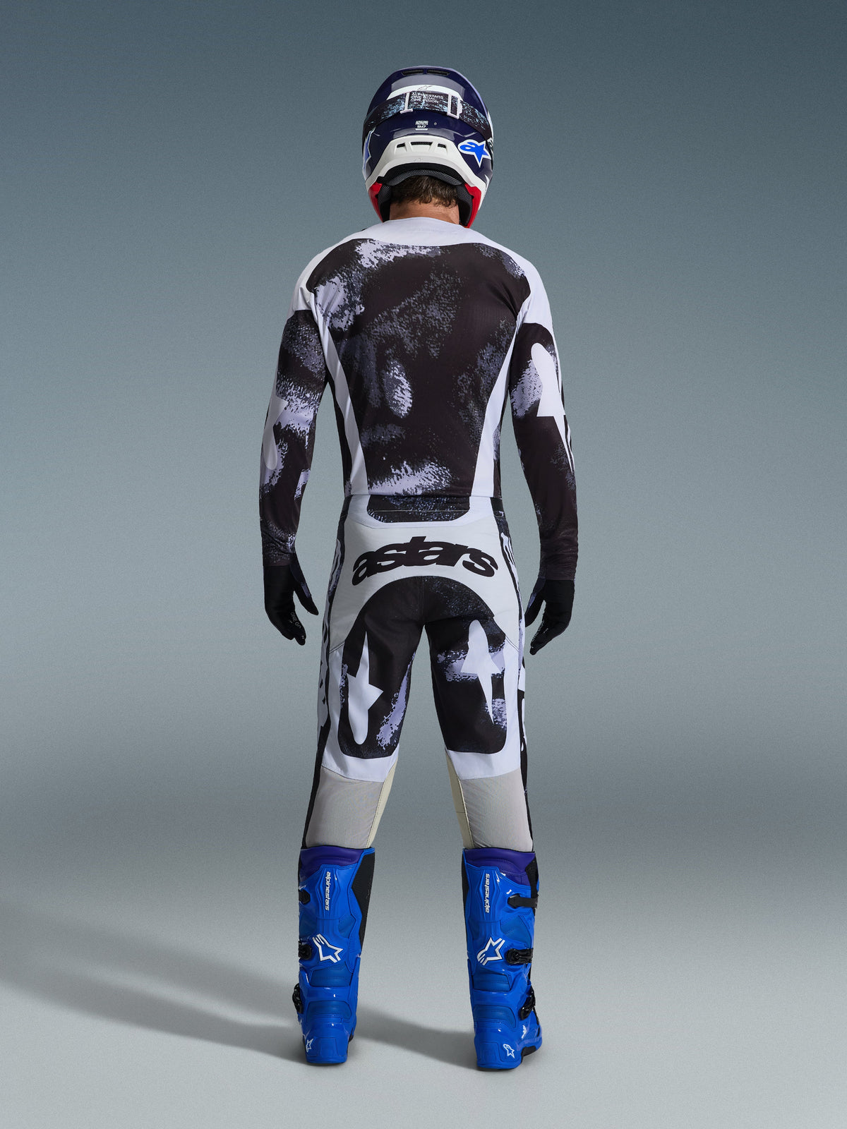 Alpinestars 2026 Racer Lahnd Pants, spodnie motocrossowe, szaro-czarny wzór Iron Camo, widok od tyłu motocyklisty w pełnym stroju wyścigowym z pasującą koszulką i niebieskimi butami, zaprojektowane z myślą o wydajności off-road