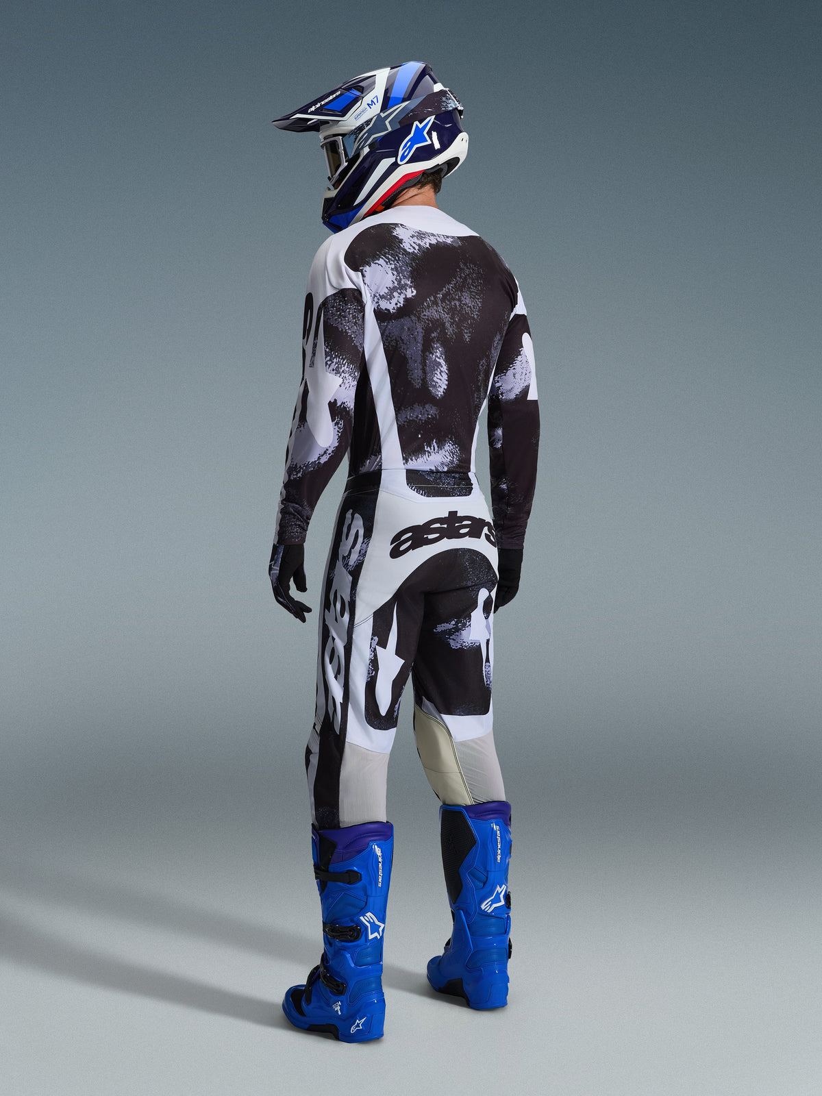 Motocyklista motocrossowy od tyłu w spodniach Alpinestars 2026 Racer Lahnd Iron Camo, dopasowana koszulka w czarno-białe wzory, jaskrawoniebieskie buty techniczne i niebiesko-biały kask, zestaw odzieży off-road z wyraźnym brandingiem 'astars' na siedzisku.