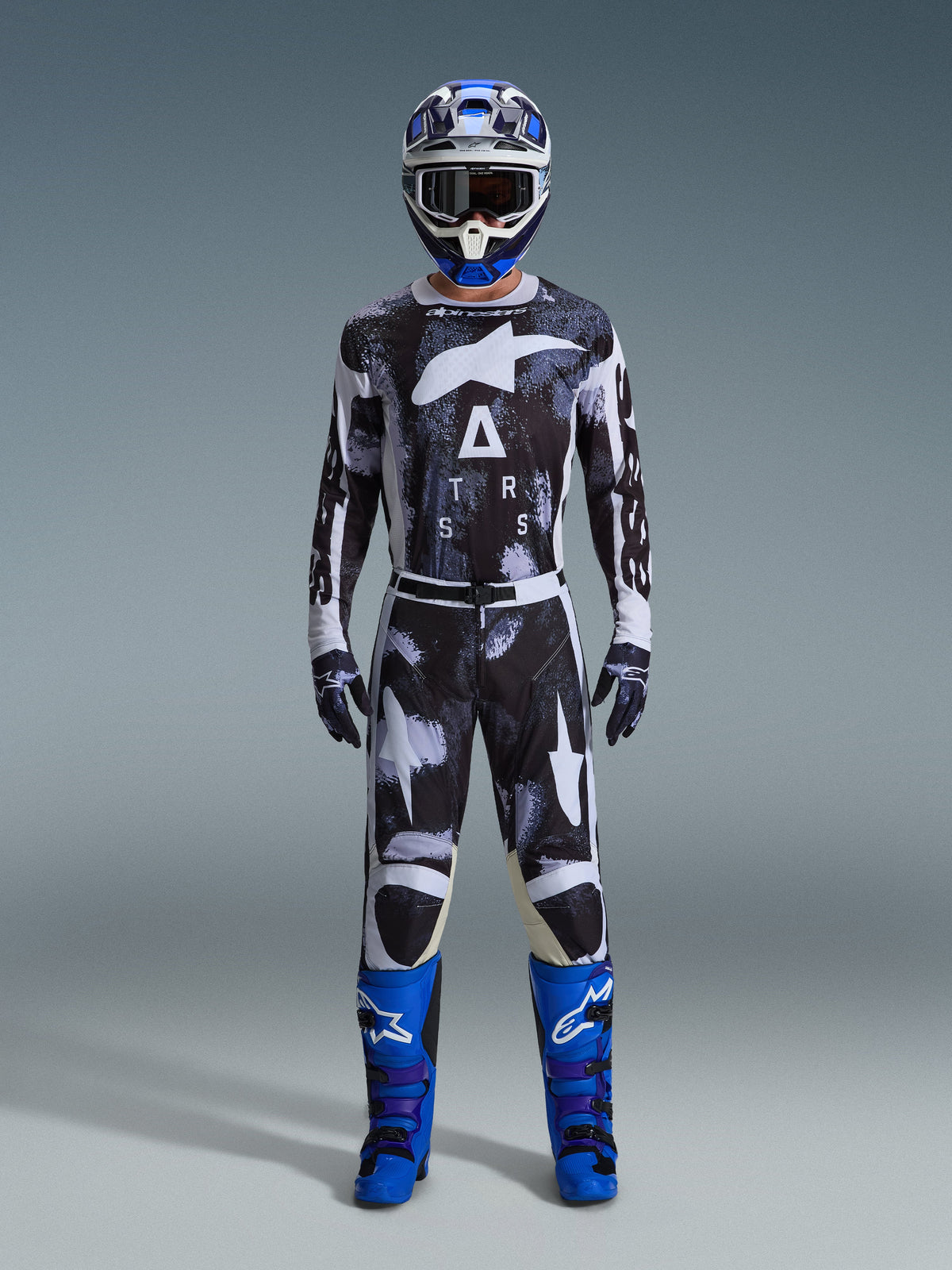 Motocyklista motocrossowy w spodniach Alpinestars 2026 Racer Lahnd w kolorze Iron Camo, szaro-czarny wzór kamuflażu, dopasowana koszulka, niebiesko-biały kask z goglami i jaskrawoniebieskie buty, kompletny zestaw odzieży motocrossowej
