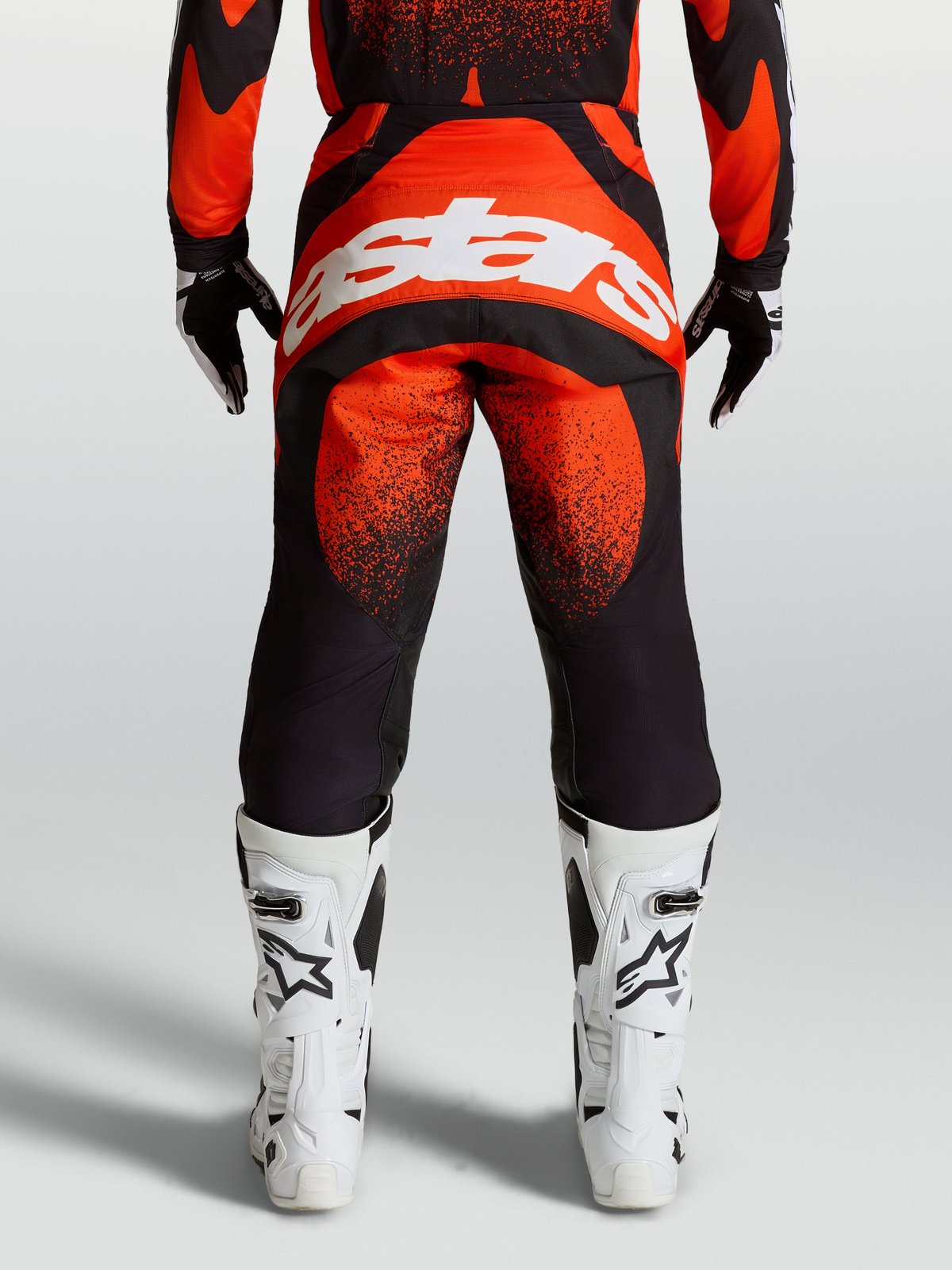 Alpinestars Spodnie Techstar Swan, spodnie motocrossowe, czerwone i czarne, widok z tyłu na modelu, białe logo Astars, białe buty wyścigowe.