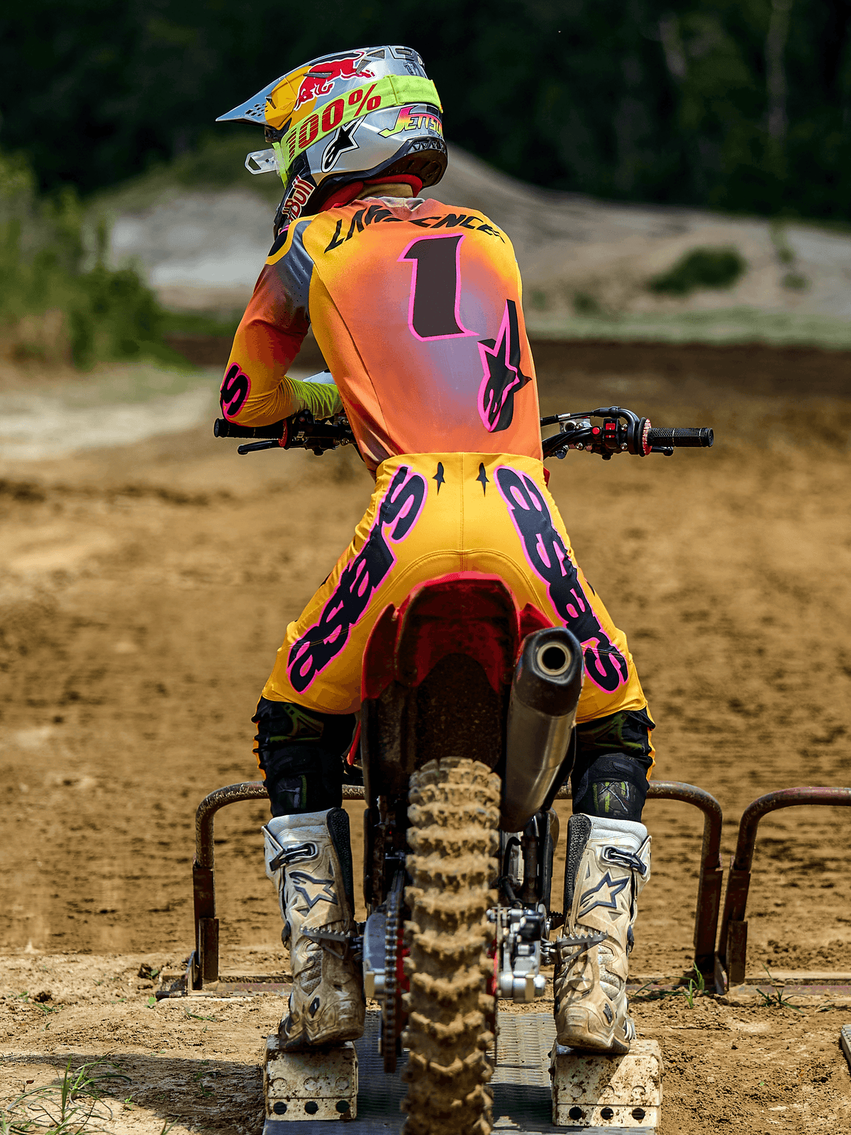 Zawodnik motocrossowy widziany z tyłu na bramce startowej, ubrany w spodnie Alpinestars 2025 Supertech Lipan w kolorze żółtym i różowym, pasującą koszulkę z różowymi akcentami i numerem 1, białe buty Alpinestars oraz kask wyścigowy, profesjonalny zestaw odzieży do wyścigów terenowych