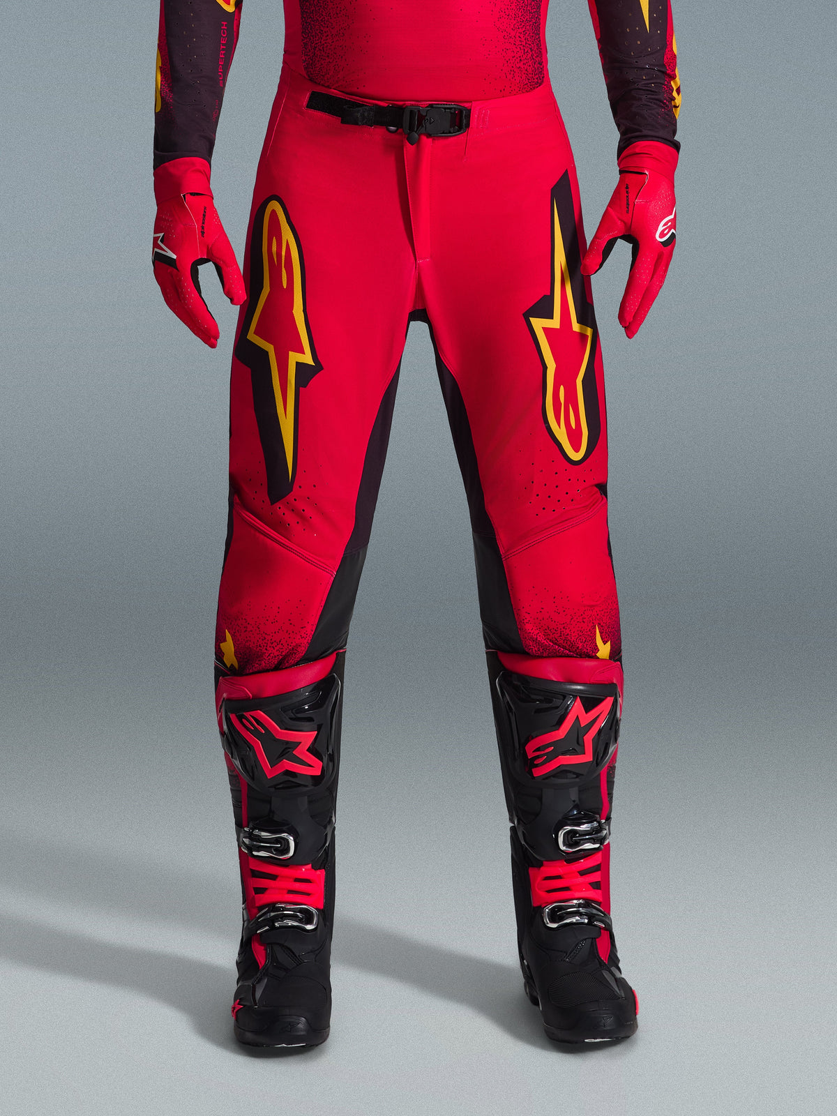 Alpinestars 2026 Supertech Scenz Pants, spodnie motocrossowe, czerwone, czarne i żółte, profesjonalny sportowy krój z dużymi żółtymi logo gwiazdy na udach, w zestawieniu z pasującymi czerwonymi rękawicami i czarno-czerwonymi butami technicznymi, zaprojektowane do wyczynowej jazdy terenowej