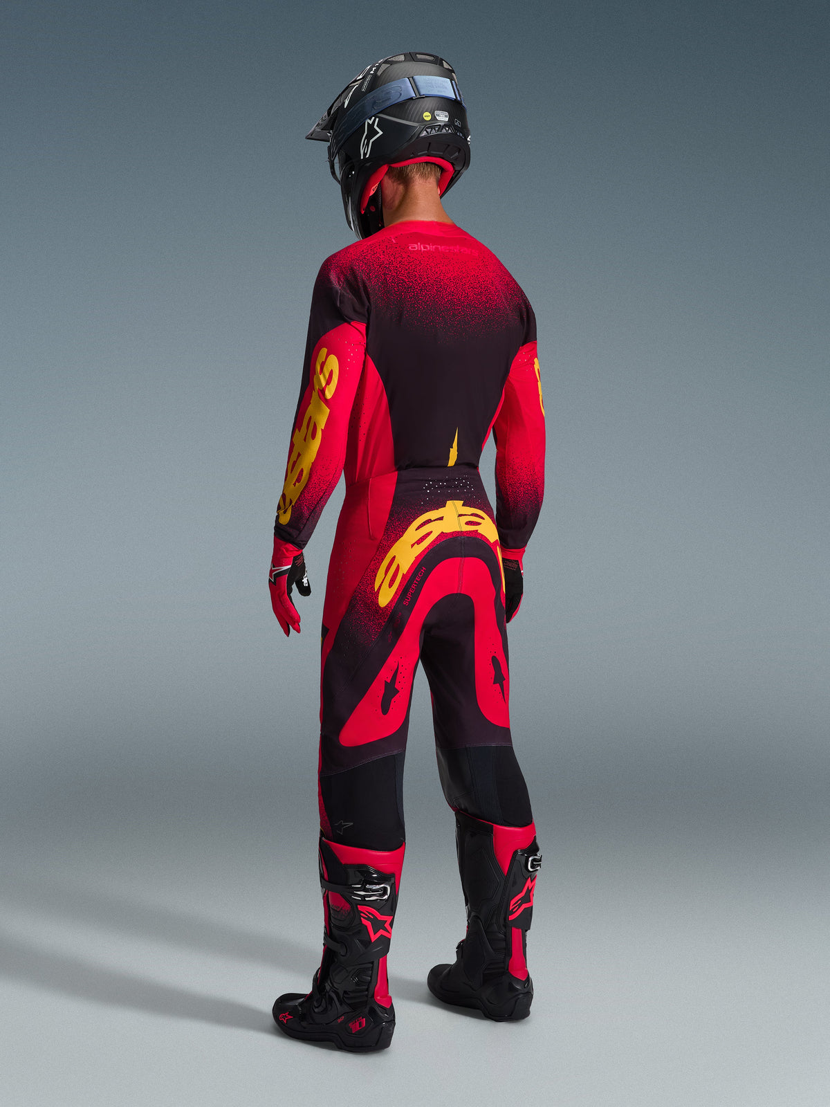 Motocyklista widziany od tyłu, ubrany w spodnie Alpinestars Supertech Scenz 2026 w kolorze czerwono-czarno-żółtym, dopasowana koszulka z wyrazistym żółtym logo, czarno-czerwone buty motocrossowe i czarny kask, kompletny profesjonalny zestaw wyścigowy do jazdy terenowej.
