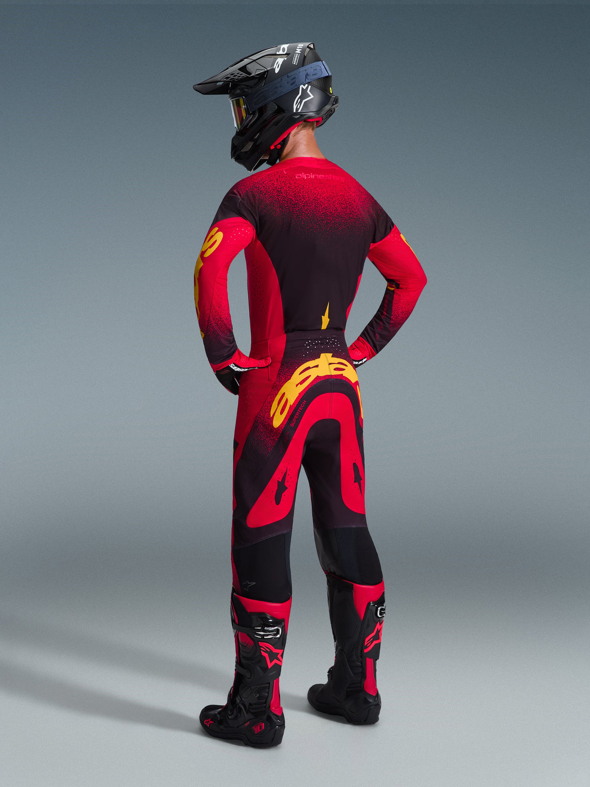 Zawodnik motocrossowy widziany od tyłu w spodniach Alpinestars 2026 Supertech Scenz w kolorze czerwono-czarno-żółtym, z dużym żółtym logo na siedzeniu, w zestawie z pasującą koszulką z czerwono-czarnym gradientem, czarnym kaskiem i czerwonymi butami motocrossowymi; profesjonalny zestaw odzieży wyścigowej do jazdy off-road.