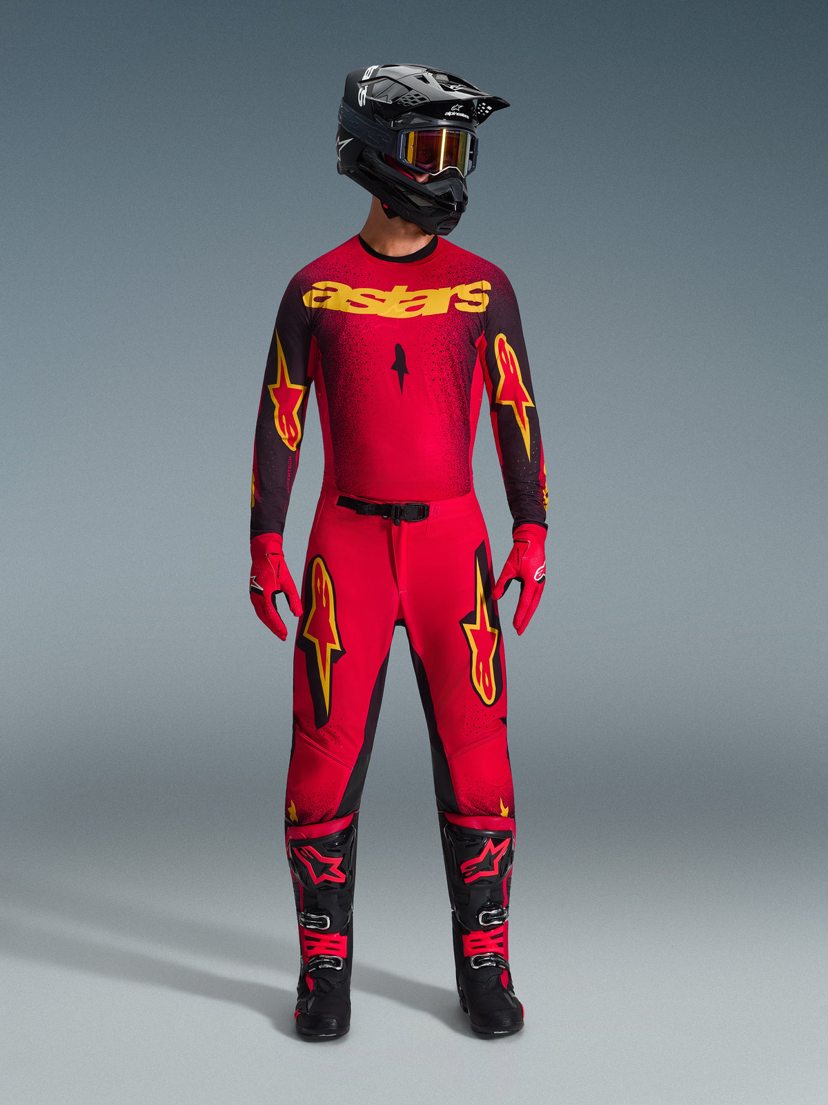 Alpinestars Supertech Scenz Pants 2026, spodnie motocrossowe, czerwone, czarne i żółte, prezentowane przez zawodnika w pełnym zestawie wyścigowym, pasująca czerwono-czarna bluza z żółtym logo, czarny kask, czerwone rękawice i czarne buty, sportowy krój pro-fit do zawodów terenowych