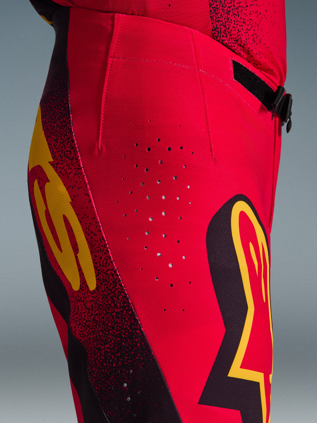 Alpinestars 2026 Supertech Scenz Pants, spodnie motocrossowe, czerwone, czarne i żółte, zbliżenie z boku na laserowo wycinane otwory wentylacyjne, wyraziste logo i elastyczną tkaninę rip-stop