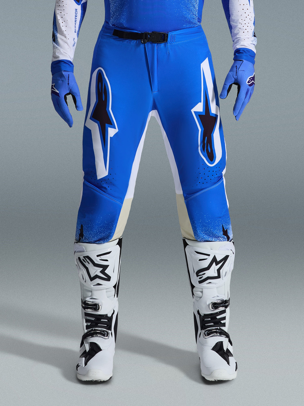 Alpinestars 2026 Supertech Scenz Pants, spodnie motocrossowe, niebiesko-białe (Ucla Blue White), prezentowane z dopasowanymi niebieskimi rękawicami i białymi butami, duże białe i czarne grafiki na udach, sportowy krój do jazdy w terenie