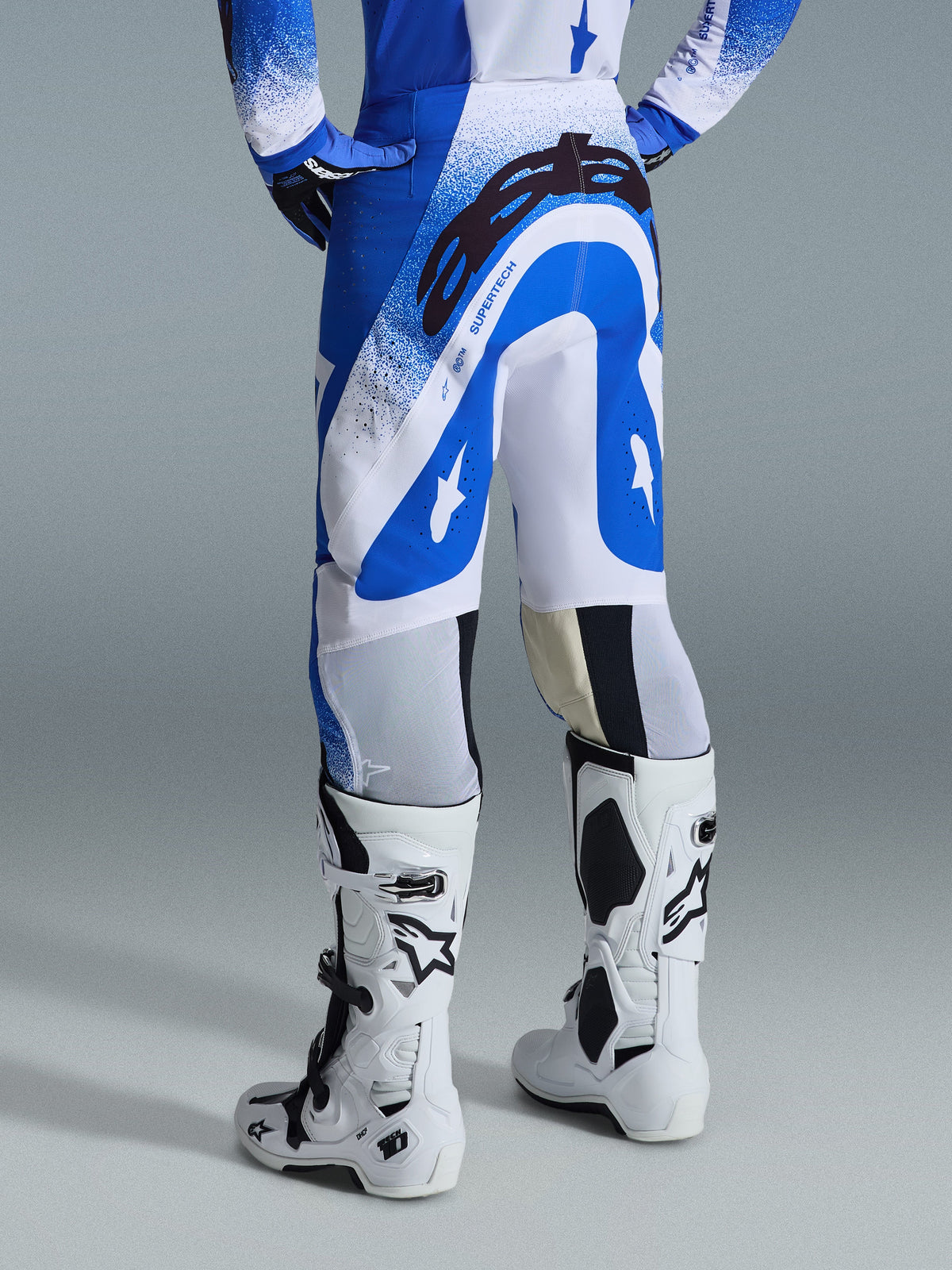 Alpinestars Supertech Scenz Pants 2026, spodnie motocrossowe, niebiesko-białe, widok z tyłu ukazujący duże czarne logo Astars na siedzeniu i branding „SUPERTECH”, zestawione z białymi butami Tech 10 oraz dopasowaną niebiesko-białą koszulką.