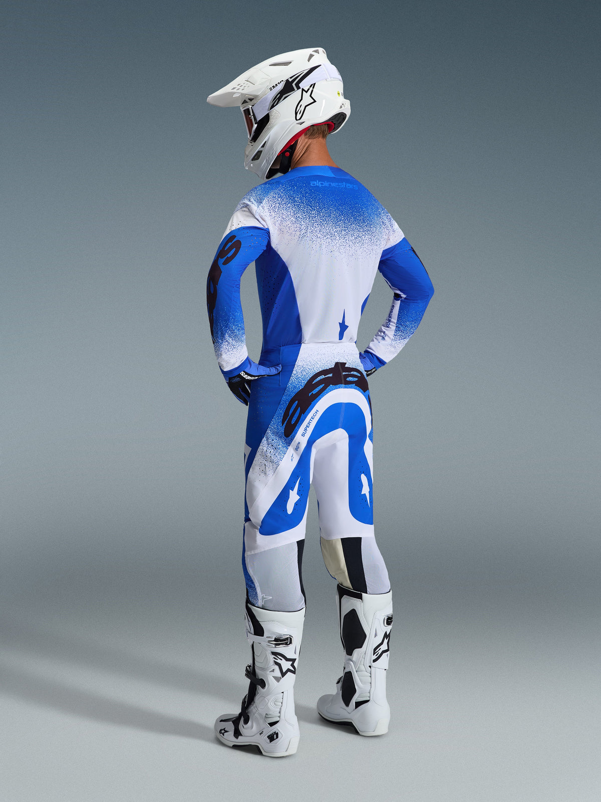 Zawodnik motocrossowy widoczny od tyłu w spodniach i koszulce 2026 Supertech Scenz w kolorystyce Ucla Blue White, biały kask z logo Alpinestars, biało-czarne buty motocrossowe, profesjonalny zestaw odzieży wyścigowej off-road