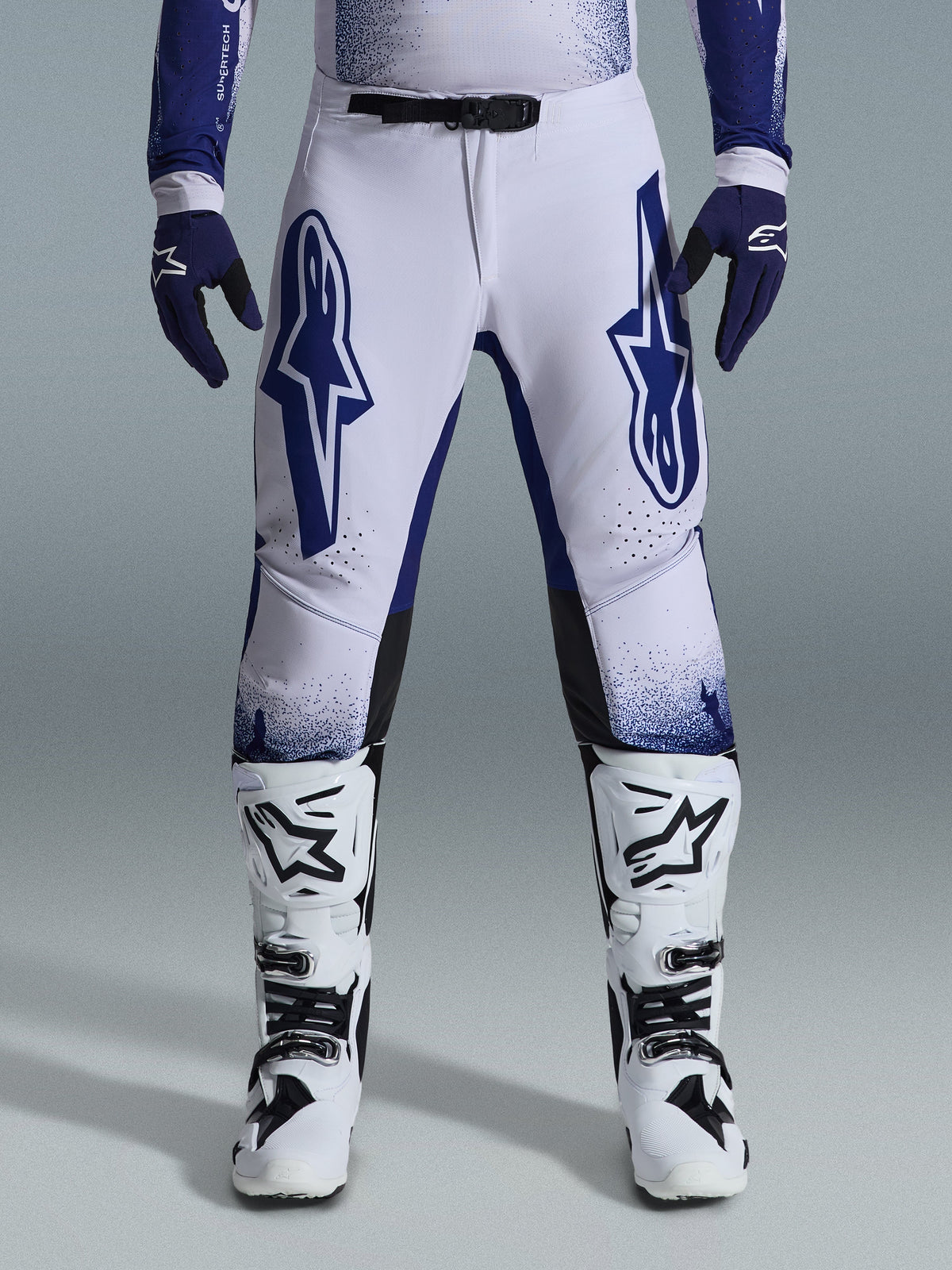 Alpinestars 2026 Supertech Scenz Pants, Motocross Pants, Light Gray and Dark Blue, worn with white and black motocross boots and dark blue gloves, featuring large blue Alpinestars logos on the thighs, SX1 magnetic buckle, and athletic fit for off-road racing