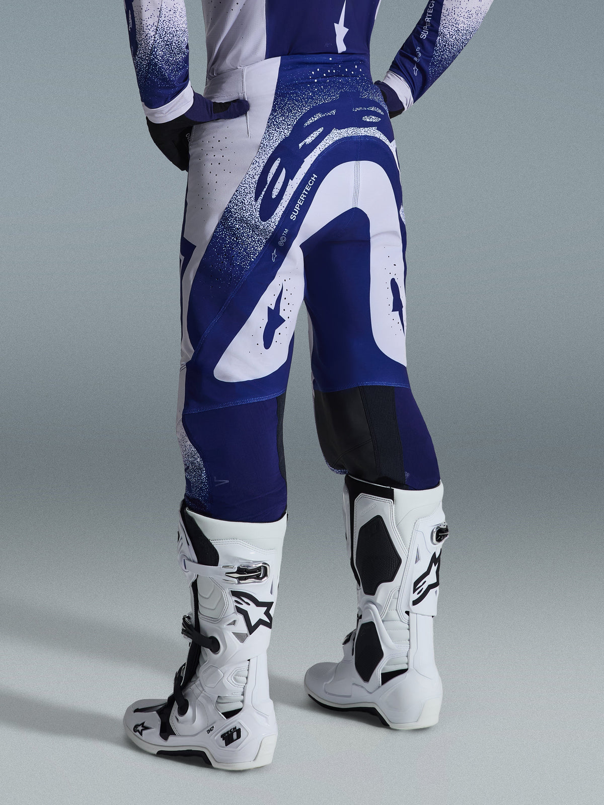 Alpinestars Supertech Scenz Pants 2026, spodnie motocrossowe, jasnoszare i ciemnoniebieskie, widok z tyłu w zestawie z pasującą bluzą i białymi butami, wyrazista niebieska grafika z nakrapianymi akcentami, sportowy krój wyczynowy do wyścigów off-road