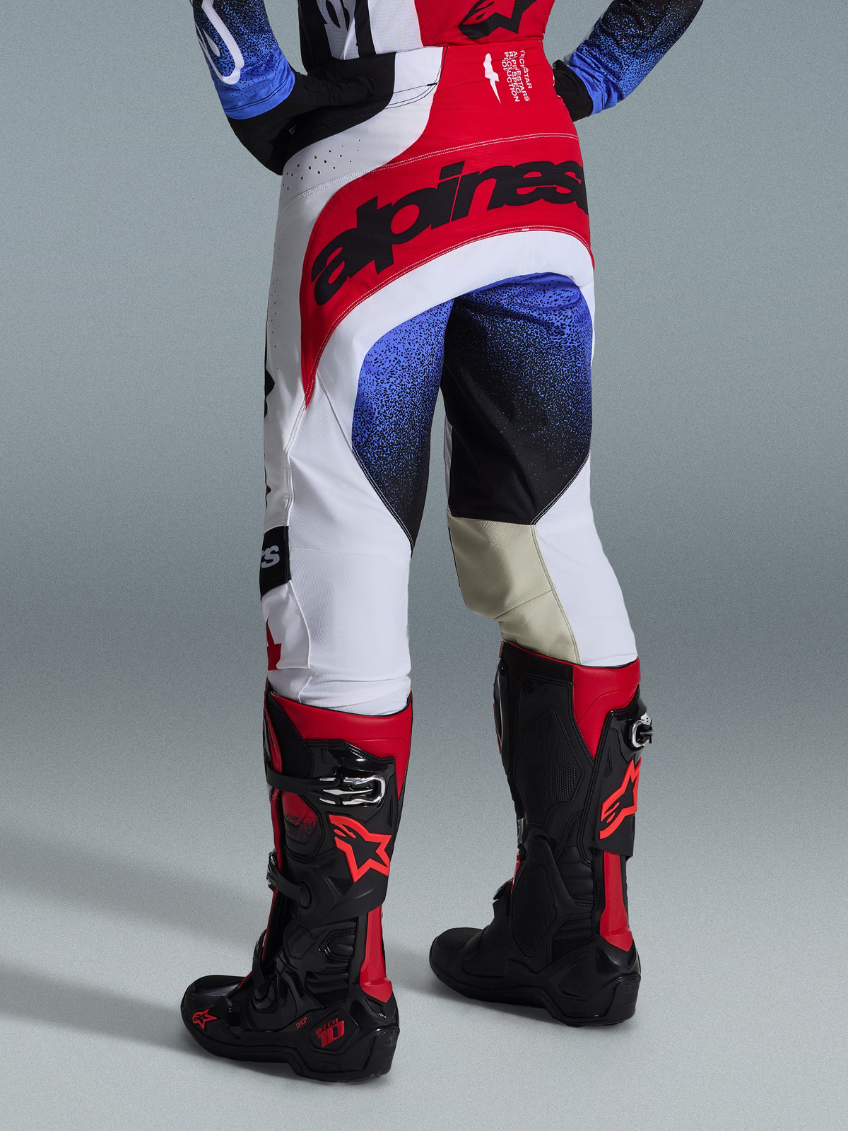 Alpinestars 2026 Techstar Knif Pants, spodnie motocrossowe, czarno-biało-czerwone z niebieskimi akcentami, widok z tyłu z wyraźnym logo w pasie i beżowymi skórzanymi panelami na wewnętrznej stronie kolan, noszone z czarno-czerwonymi butami, wyczynowa odzież wyścigowa do zawodów off-road
