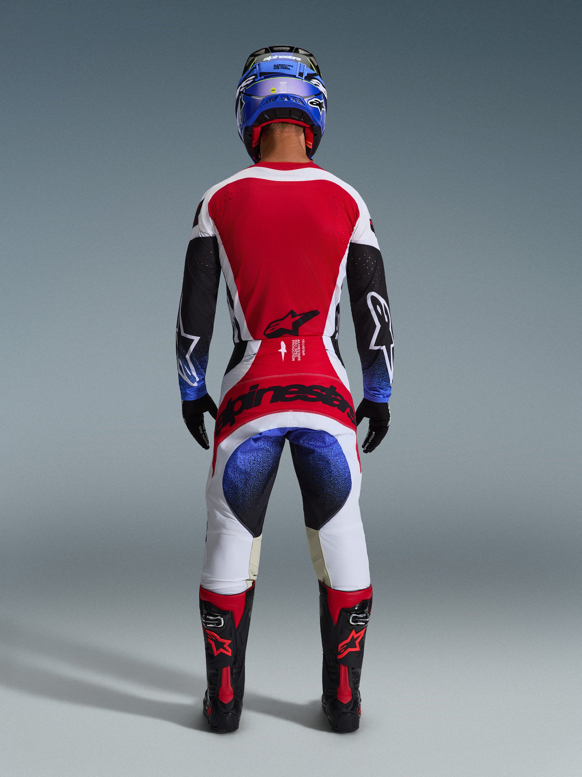 Alpinestars 2026 Techstar Knif Pants, spodnie motocrossowe, czarne, białe, czerwone i niebieskie, widok od tyłu zawodnika w pełnym stroju, w tym pasująca koszulka, niebieski kask oraz czarno-czerwone buty, z wyraźnym brandingiem Alpinestars na siedzeniu.