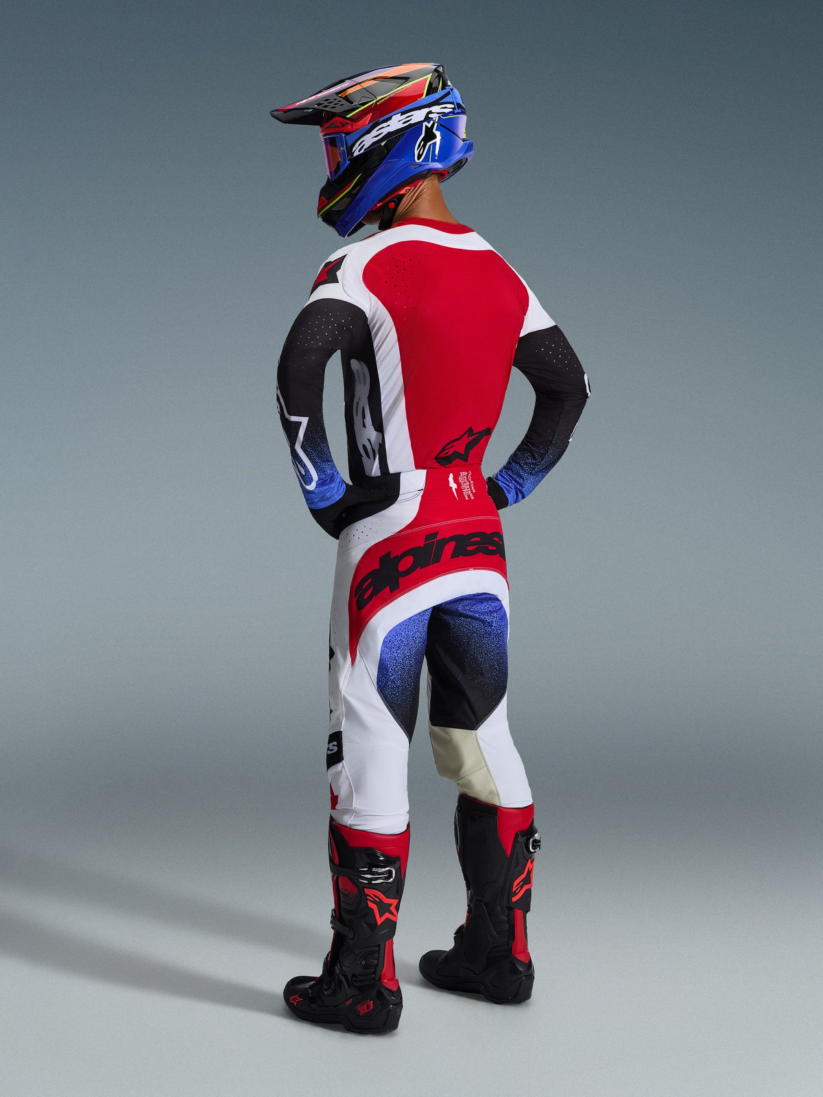 Alpinestars 2026 Techstar Knif Pants, spodnie motocrossowe, czarno-biało-czerwone z niebieskimi akcentami gradientowymi, widok od tyłu na zawodnika w pełnym stroju wyścigowym, wyraźne czerwone logo na siedzeniu, białe i czarne panele, zestawione z pasującą koszulką i butami Tech 10