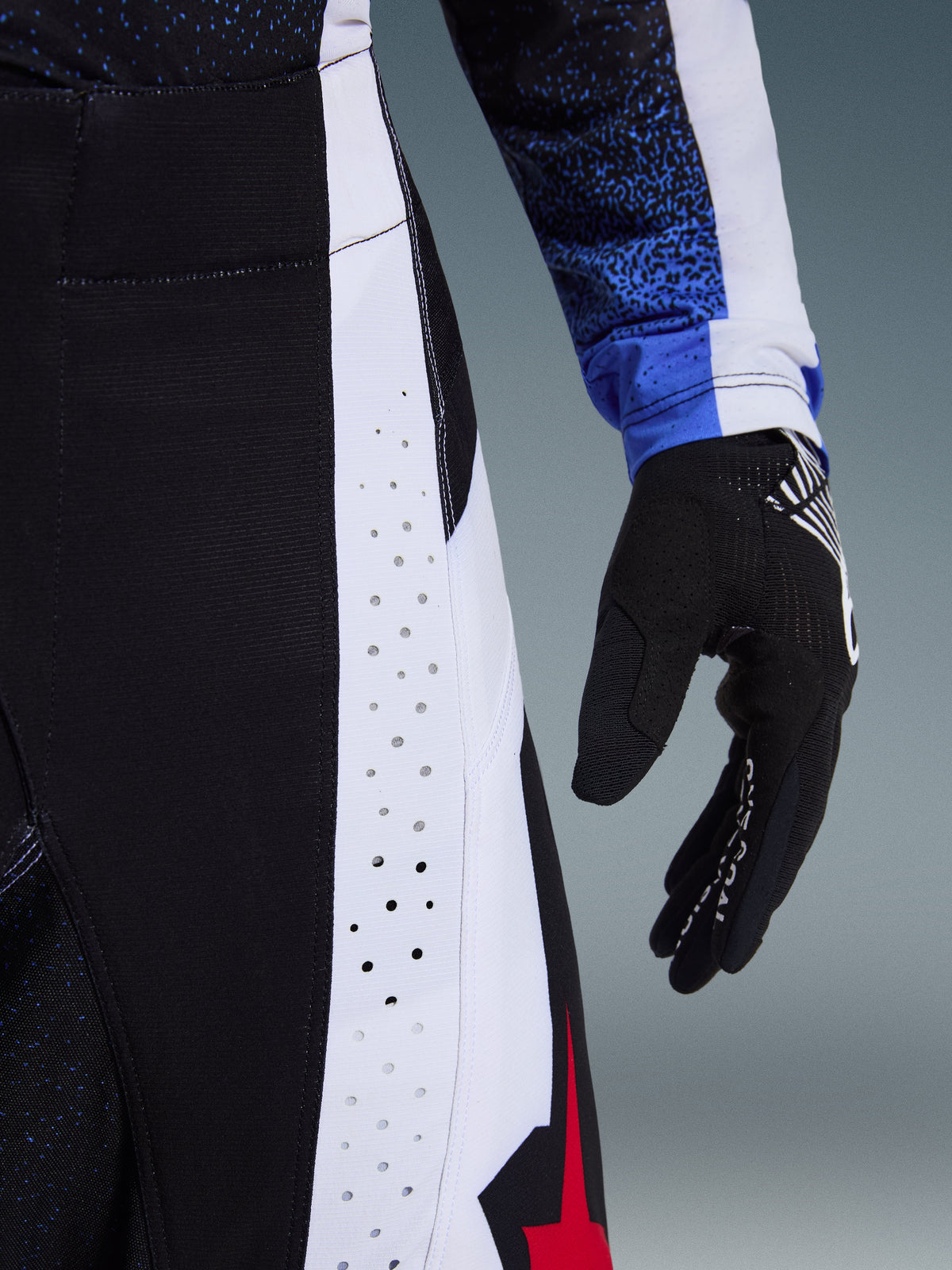 Alpinestars Techstar Knif Pants 2026, spodnie motocrossowe, czarne, białe i czerwone, zbliżenie na laserowo wycinane perforacje na panelach bocznych oraz czerwone logo z gwiazdą, zestawione z pasującą koszulką i czarną rękawicą motocrossową.