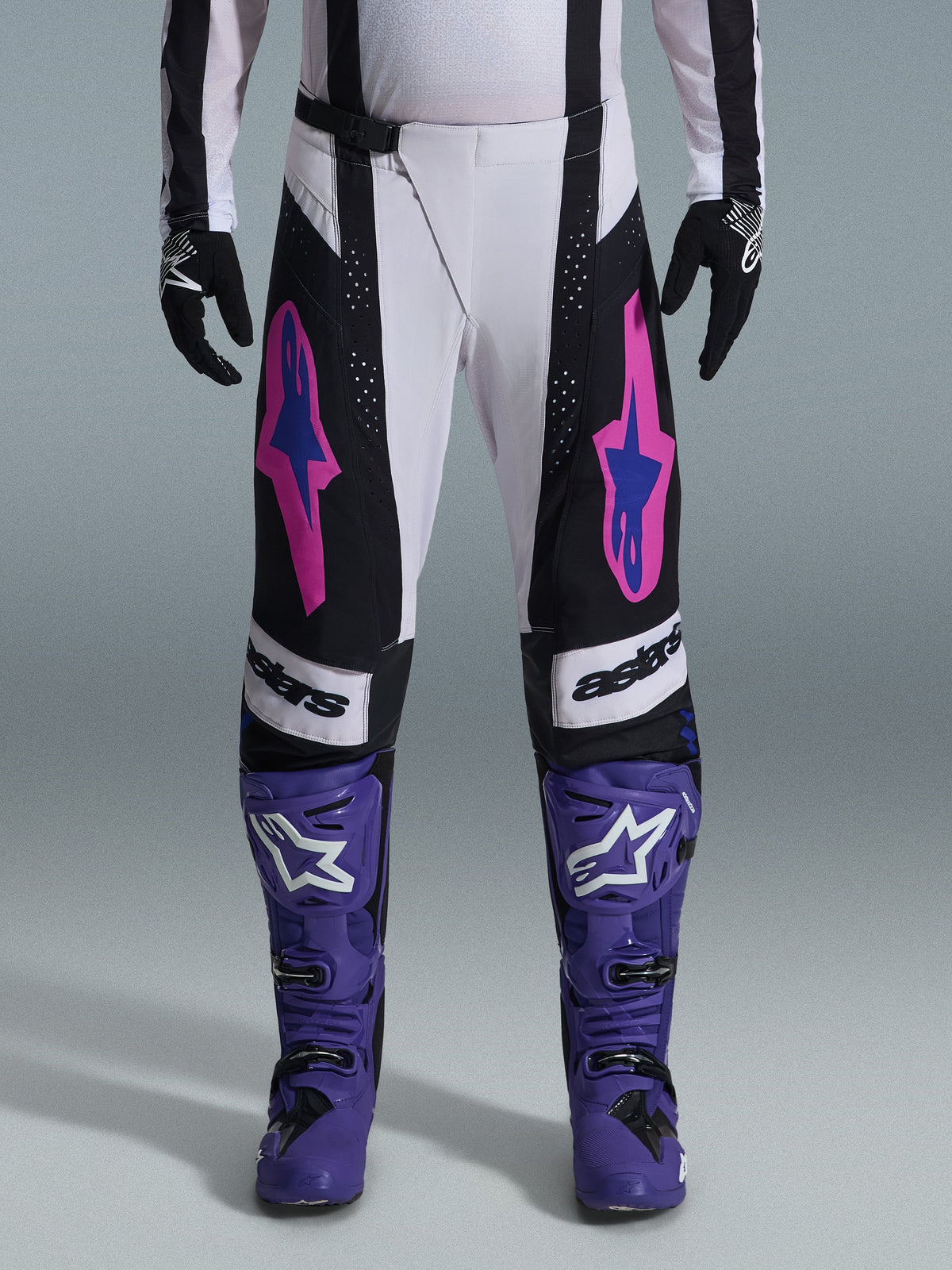 Alpinestars 2026 Techstar Knif Pants, spodnie motocrossowe, czarne, białe i fioletowe z różowymi i niebieskimi akcentami, noszone z fioletowymi butami wyścigowymi i czarnymi rękawicami, charakteryzujące się ergonomicznym krojem wyścigowym i wyrazistymi grafikami logo dla wydajności w terenie