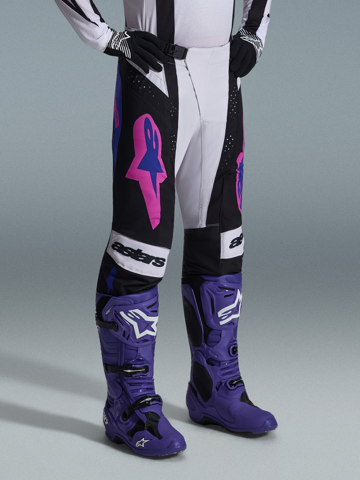 Alpinestars Techstar Knif Pants 2026, spodnie motocrossowe, czarne, jasnoszare i fioletowe, noszone z pasującymi fioletowymi butami motocrossowymi, z wyraźnymi różowymi akcentami i brandingiem „astars”, profesjonalna odzież wyścigowa