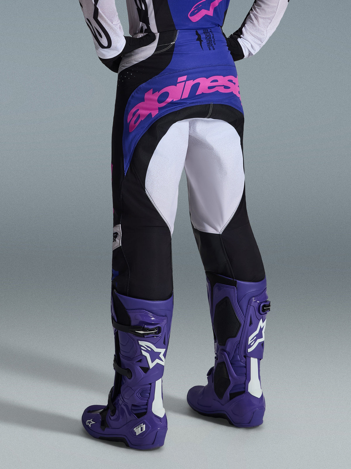 Spodnie Alpinestars Techstar Knif 2026, spodnie motocrossowe, czarne, jasnoszare i fioletowe, widok z tyłu z różowym logotypem na siedzeniu, noszone z dopasowaną koszulką i fioletowymi butami motocrossowymi, profesjonalny strój wyścigowy off-road
