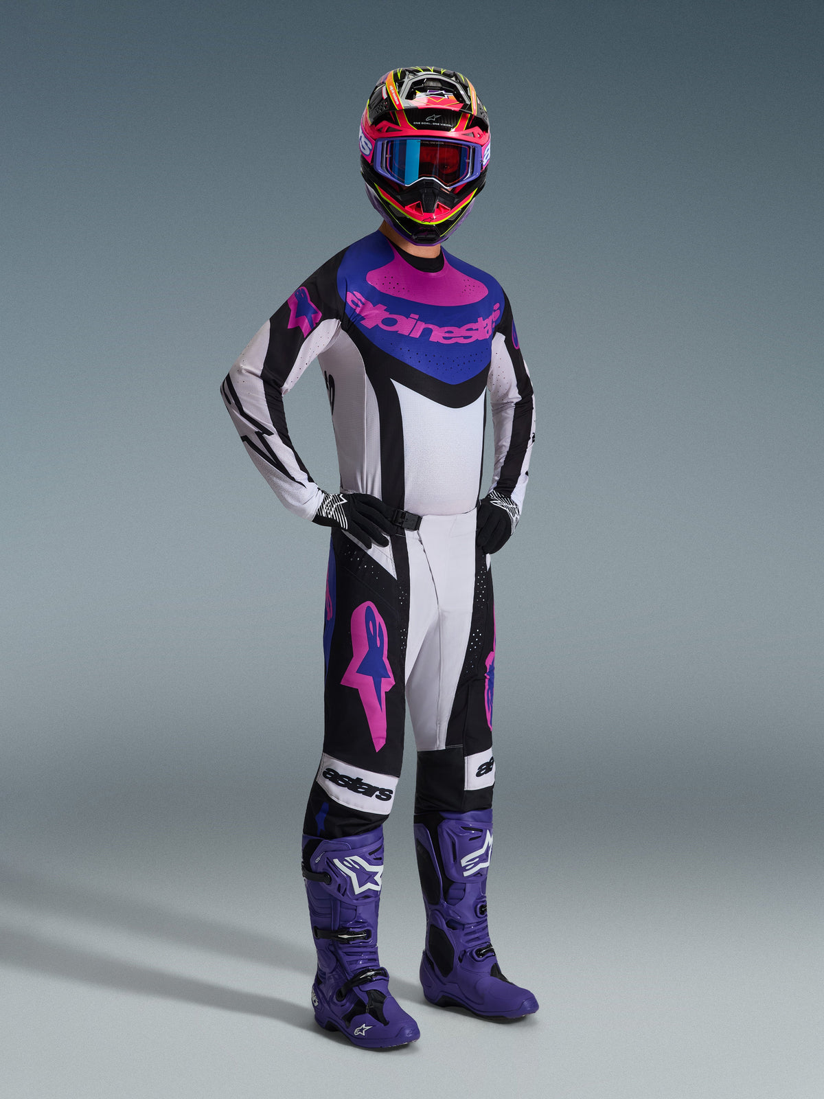 Zawodnik motocrossowy w zestawie Alpinestars 2026 Techstar Knif, biała koszulka z czarnymi, niebieskimi i różowymi akcentami, dopasowane spodnie z różowymi logotypami Astars, wielobarwny kask z goglami, fioletowe buty wyścigowe, profesjonalna odzież wyczynowa do motocrossu