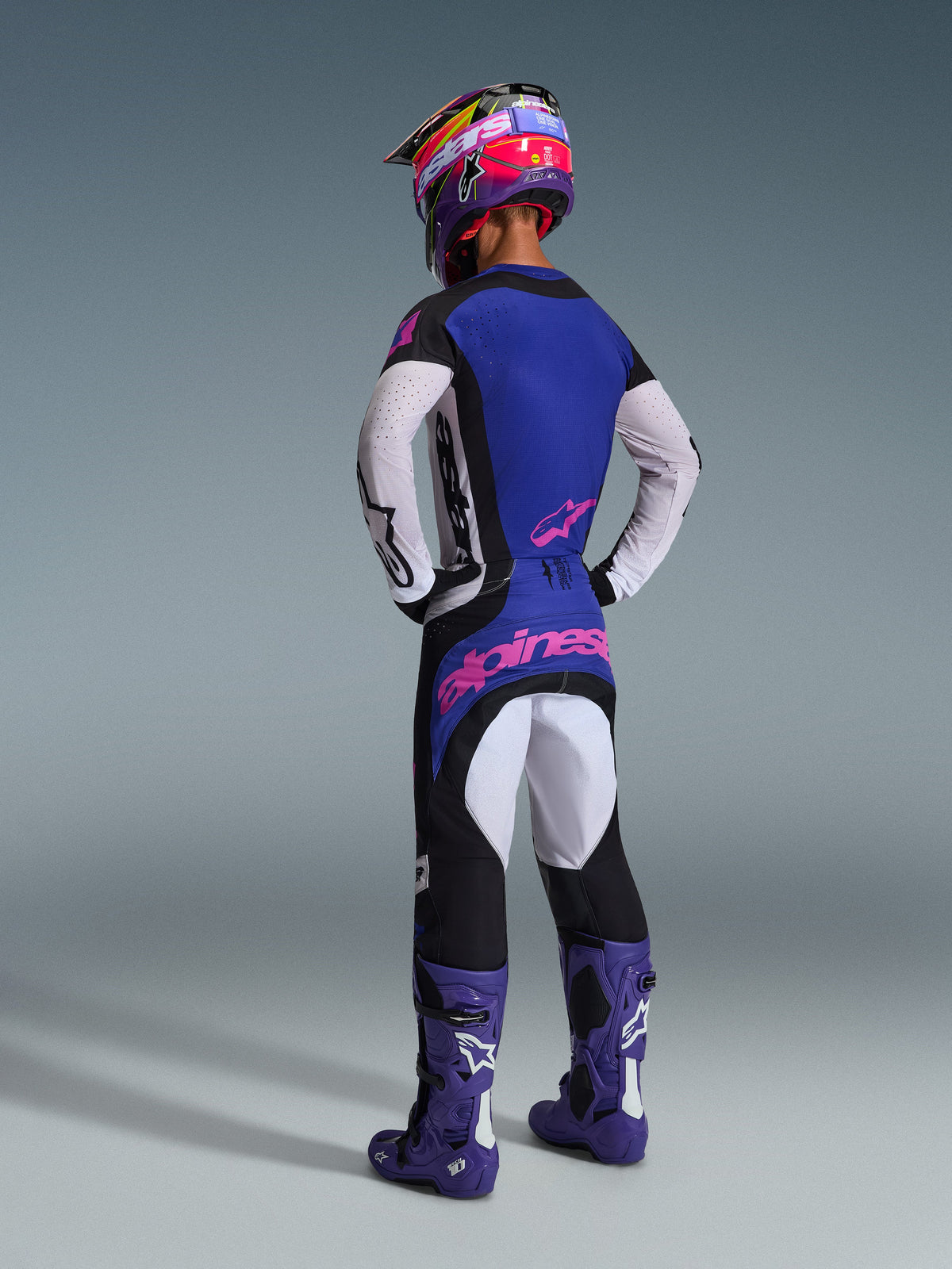 Zawodnik motocrossowy widoczny od tyłu w zestawie Alpinestars Techstar Knif 2026, niebieska koszulka z białymi perforowanymi rękawami i różowymi logo, czarno-białe spodnie z różowymi oznaczeniami marki z tyłu, fioletowe buty i jaskrawy fioletowo-różowy kask, profesjonalny strój wyścigowy.
