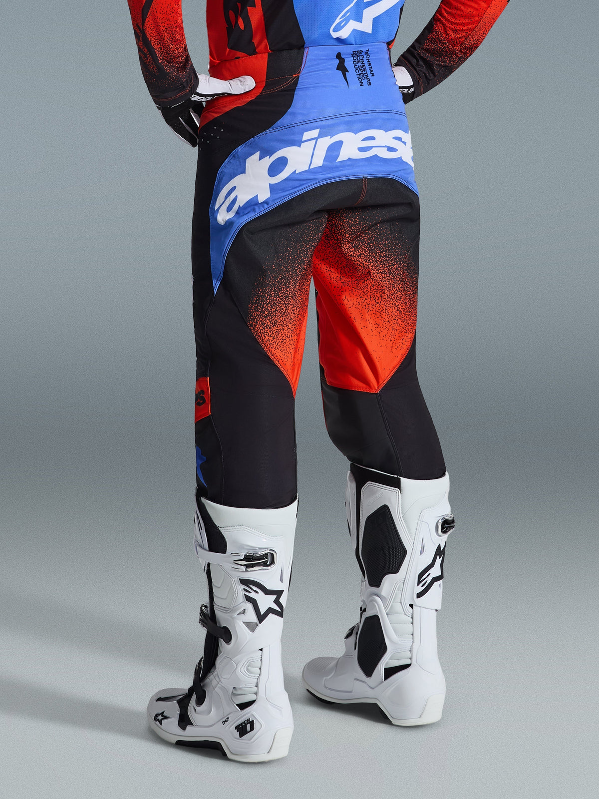 Spodnie Alpinestars 2026 Techstar Knif, spodnie motocrossowe, pomarańczowo-czarno-niebieskie, widok z tyłu pokazujący niebieski karczek z białym logo Alpinestars, pomarańczowy wzór gradientowy na nogawkach, noszone z pasującą koszulką i białymi butami do wyścigów terenowych.