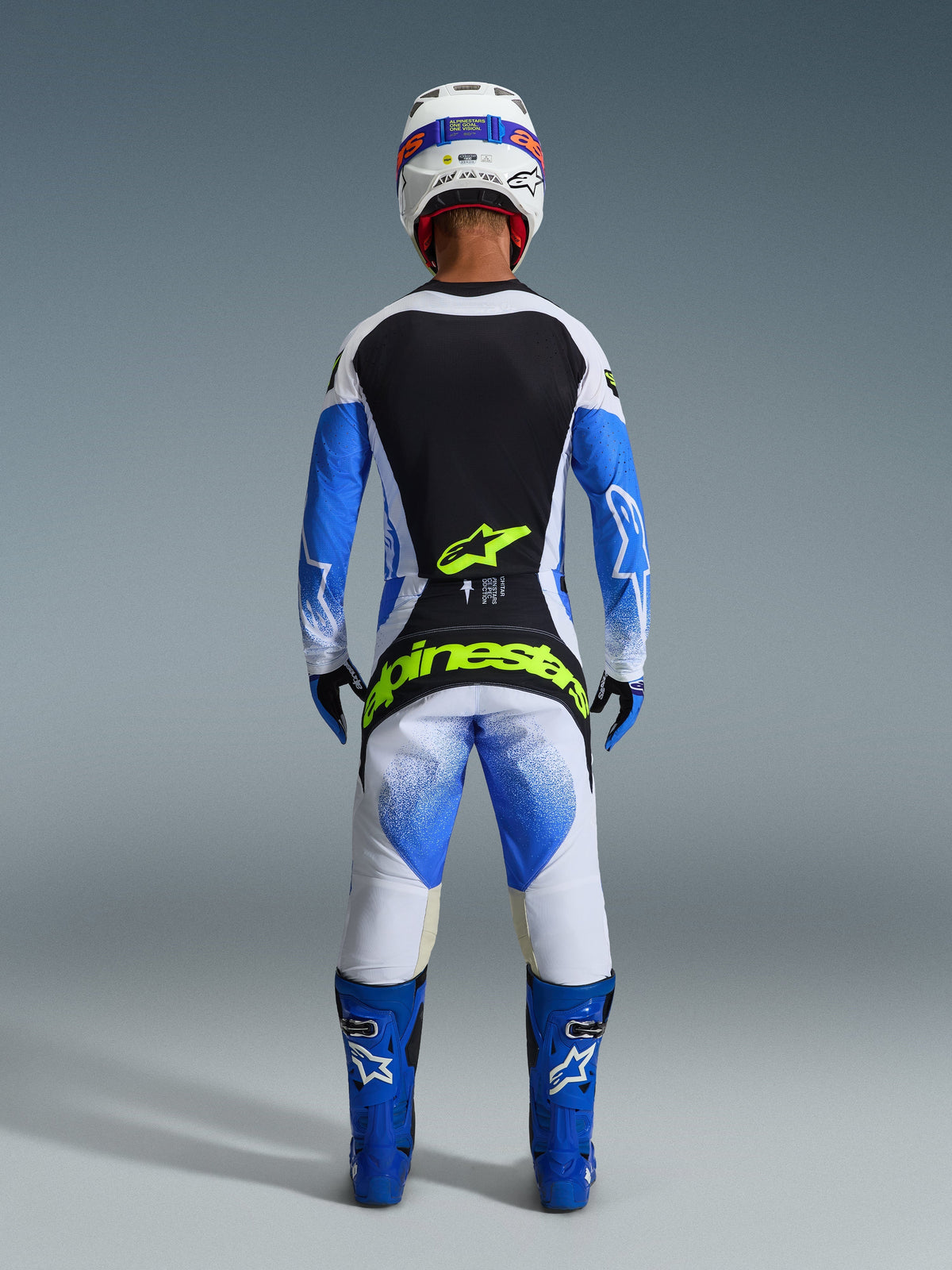Motocross rider from behind wearing complete racing gear, 2026 Techstar Knif Pants in UCLA Blue Black and Yellow Fluo, matching long-sleeve jersey with black and white panels, blue off-road boots, white helmet, featuring bold fluorescent yellow Alpinestars branding on the seat.
