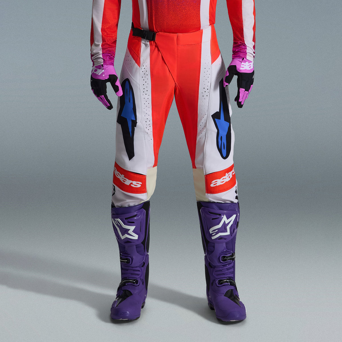 Alpinestars 2026 Techstar Knif Gear Set, dolna część ciała zawodnika motocrossowego w czerwono-białych spodniach z niebieskimi akcentami, jaskrawofioletowe buty wyścigowe z białym logo, dopasowane czarno-fioletowe rękawice, profesjonalny zestaw do wyścigów terenowych