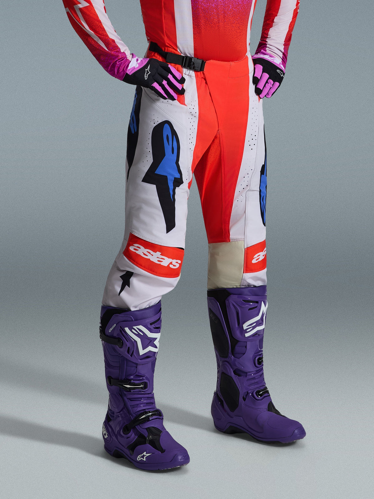 Alpinestars 2026 Techstar Knif Pants, spodnie motocrossowe, biało-pomarańczowe z niebieskimi akcentami, element kompletnego zestawu z czerwono-różową koszulką, fioletowymi butami i różowymi rękawicami, wyraziste logotypy, sportowy krój wyścigowy