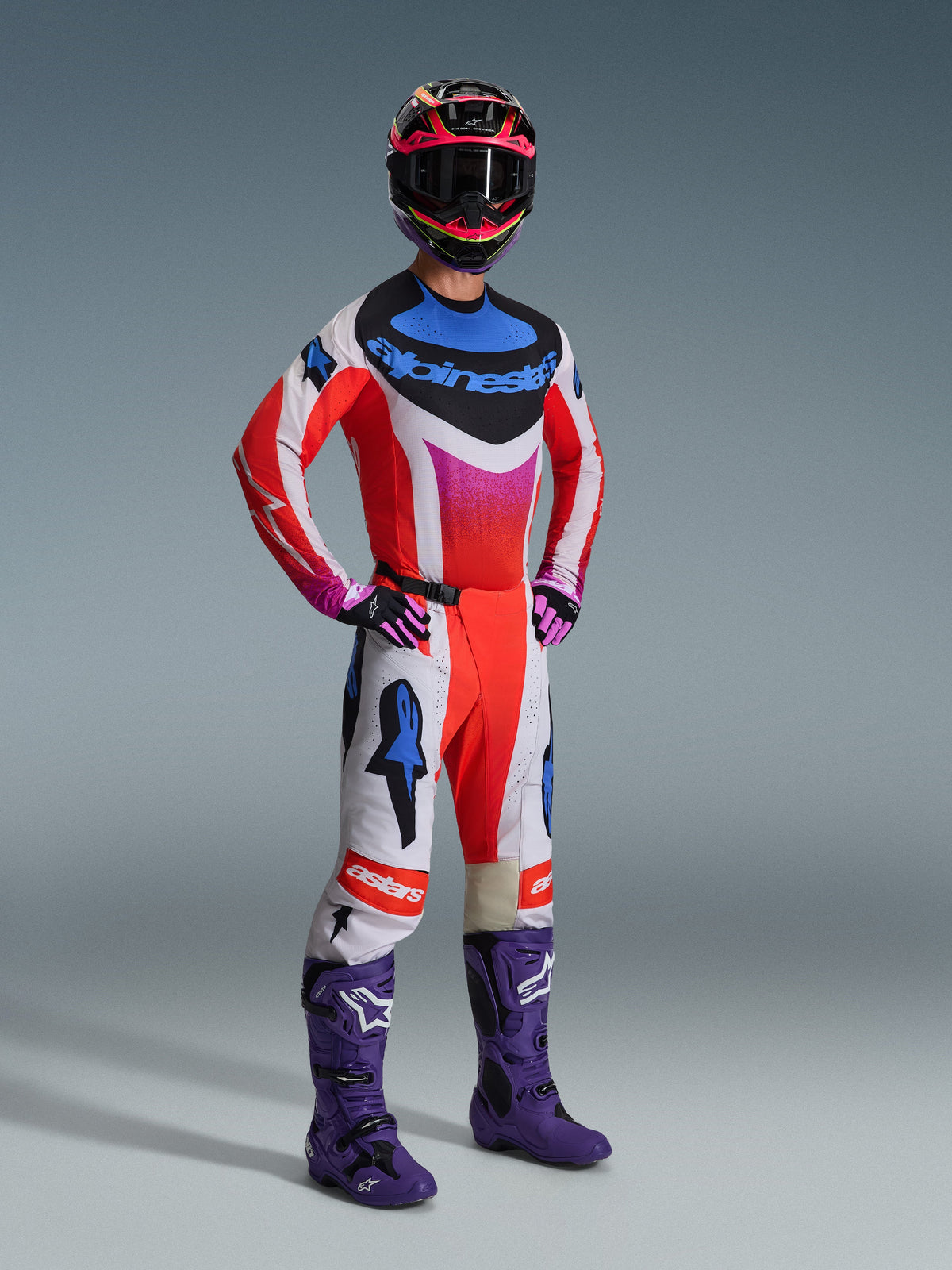 Zestaw Alpinestars 2026 Techstar Knif, motocyklista motocrossowy w pełnym stroju wyścigowym, czerwona koszulka z białymi i niebieskimi akcentami, białe spodnie z czerwonymi i niebieskimi logotypami gwiazdy, fioletowe buty motocrossowe, czarno-różowy kask, profesjonalny zestaw wyścigowy off-road