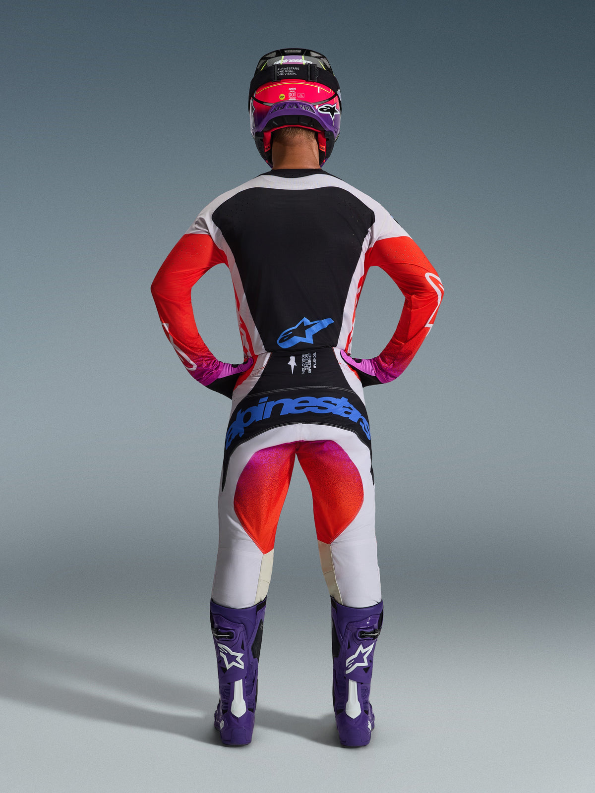 Alpinestars 2026 Techstar Knif Gear Set, kompletny strój motocrossowy widoczny od tyłu, biało-czarna koszulka z czerwonym gradientem na rękawach, białe spodnie z czerwonymi i różowymi akcentami, fioletowe buty i różowy kask, wyraźny niebieski branding, profesjonalny zestaw wyścigowy off-road