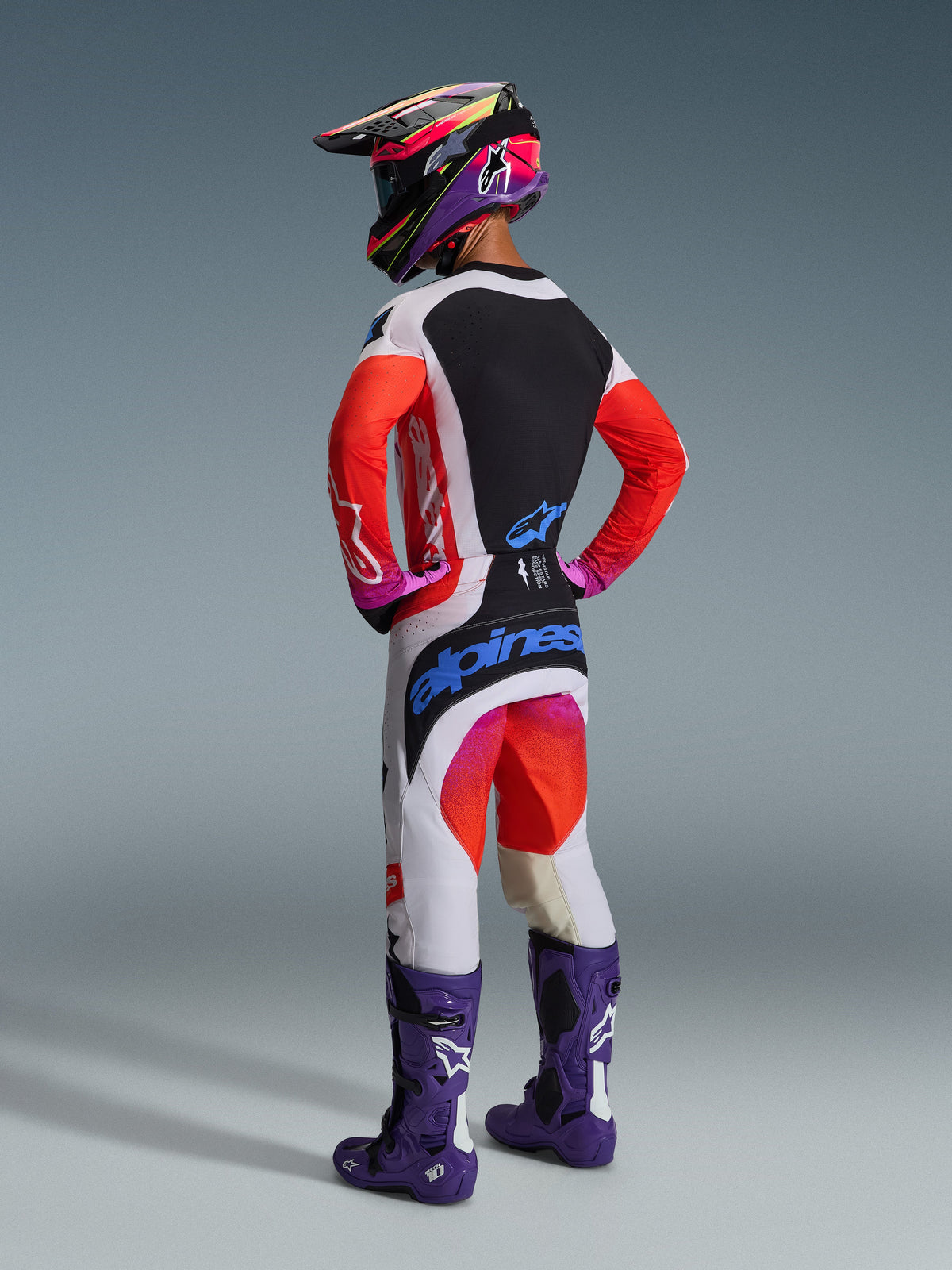 Motocyklista motocrossowy widziany od tyłu w zestawie Alpinestars 2026 Techstar Knif, biała koszulka z czerwonymi rękawami i czarnym panelem, białe spodnie z czerwonymi i fioletowymi akcentami oraz niebieskim logo Alpinestars, fioletowe buty wyścigowe i pasujący kask, profesjonalny zestaw do wyścigów terenowych
