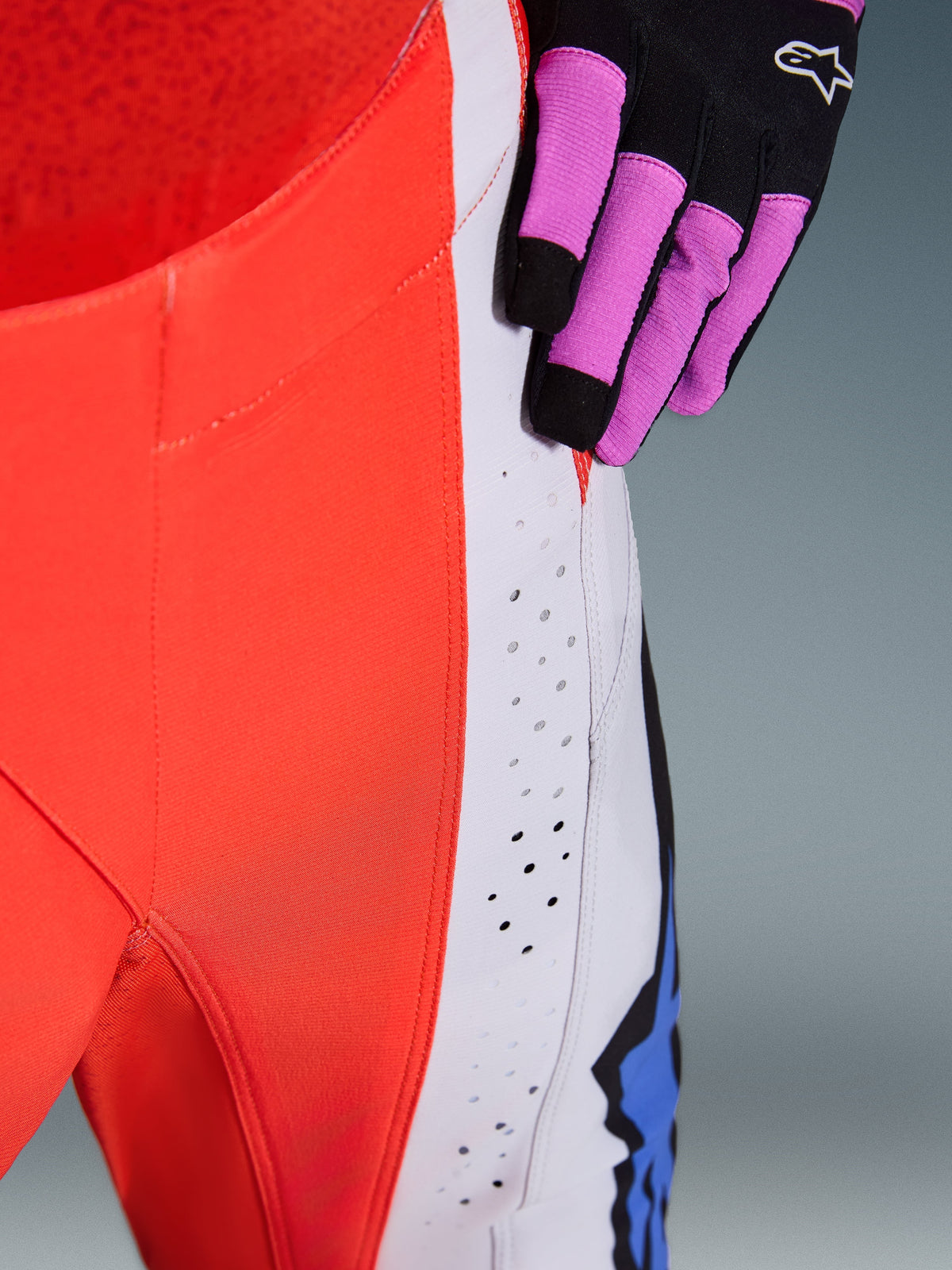 Spodnie Alpinestars Techstar Knif 2026, zbliżenie na biodro i panel boczny w kolorach pomarańczowym fluo i jasnoszarym, laserowo wycinane perforacje zapewniające przepływ powietrza, widoczna czarno-fioletowa rękawica motocrossowa, wysokiej klasy odzież wyścigowa