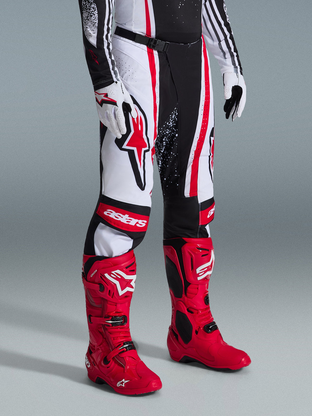 Alpinestars Techstar Nomur Pants 2026, spodnie motocrossowe, czarno-biało-czerwone, zestawione z jaskrawoczerwonymi butami wyścigowymi i białymi rękawicami, wyraźne pionowe pasy, duże czerwone logo gwiazdy na udzie, wzmocnione panele na kolanach.