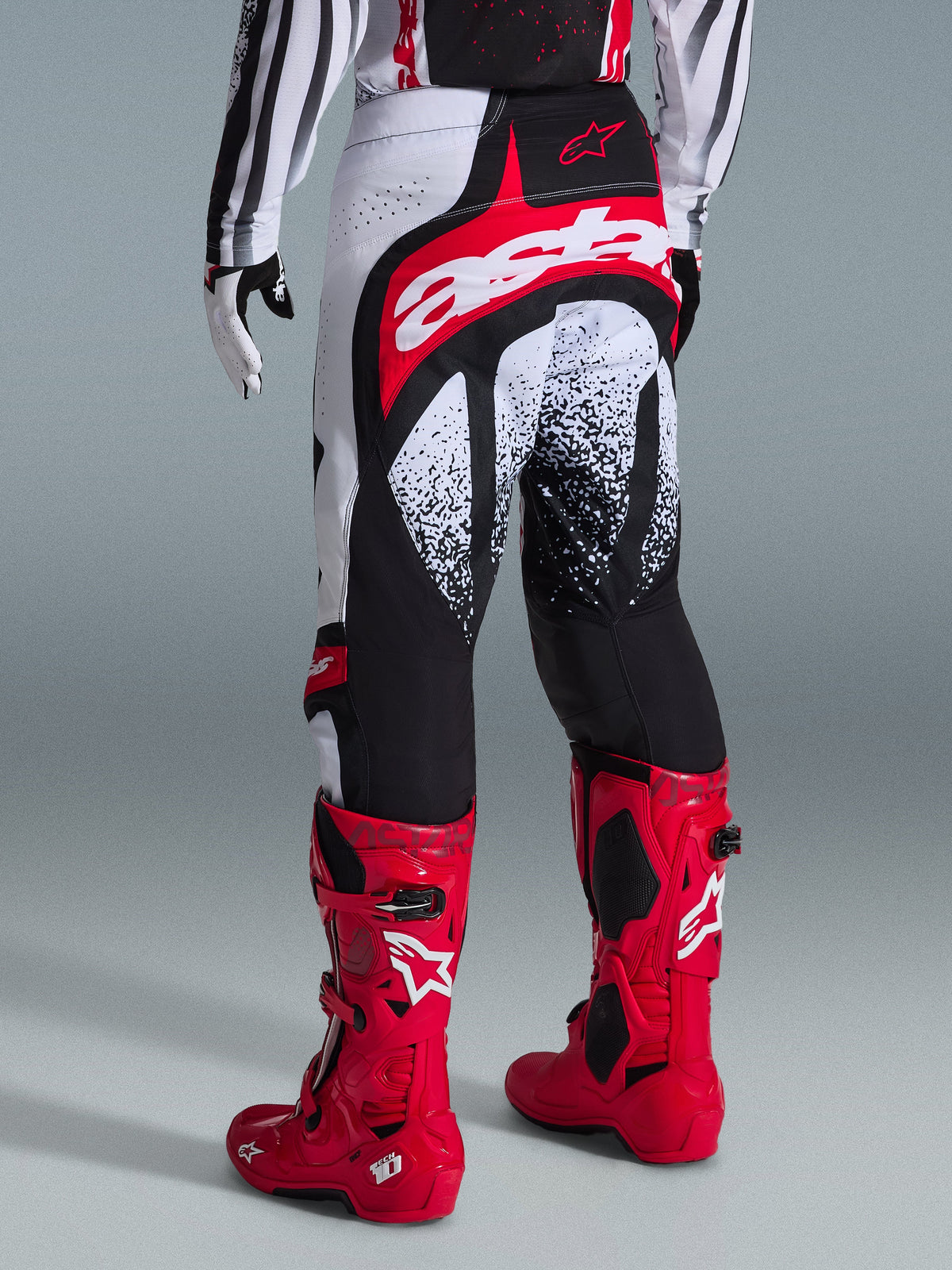 Alpinestars Techstar Nomur Pants 2026, spodnie motocrossowe, czarno-biało-czerwone, widok z tyłu na motocykliście w czerwonych butach, charakterystyczne logo „astars” na siedzeniu i nakrapiane wzory, profesjonalny sprzęt off-road