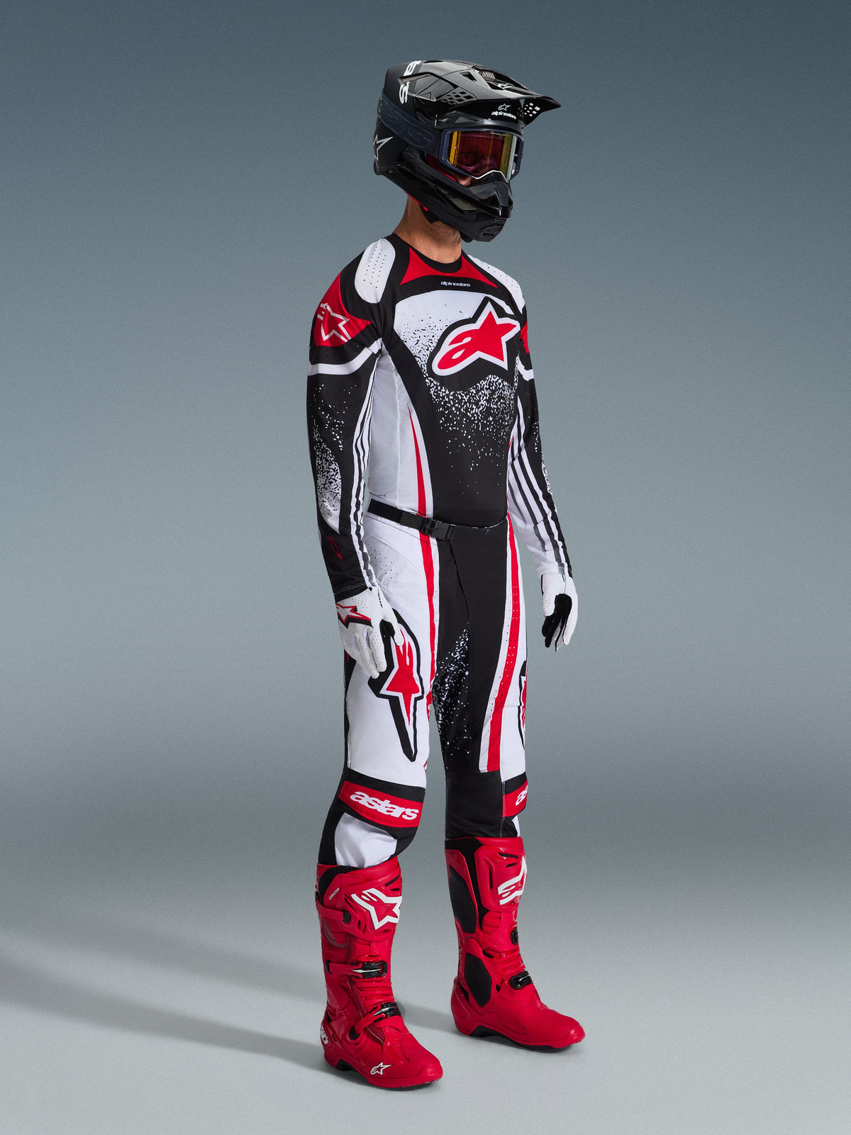 Zawodnik motocrossu w kompletnym stroju wyścigowym, spodnie 2026 Techstar Nomur w kolorze czarno-biało-czerwonym, pasująca koszulka z wyraźnym logo Alpinestars i nakrapianym wzorem, jaskrawoczerwone buty motocrossowe, biało-czarne rękawice oraz czarny kask z goglami, profesjonalny zestaw do jazdy off-road