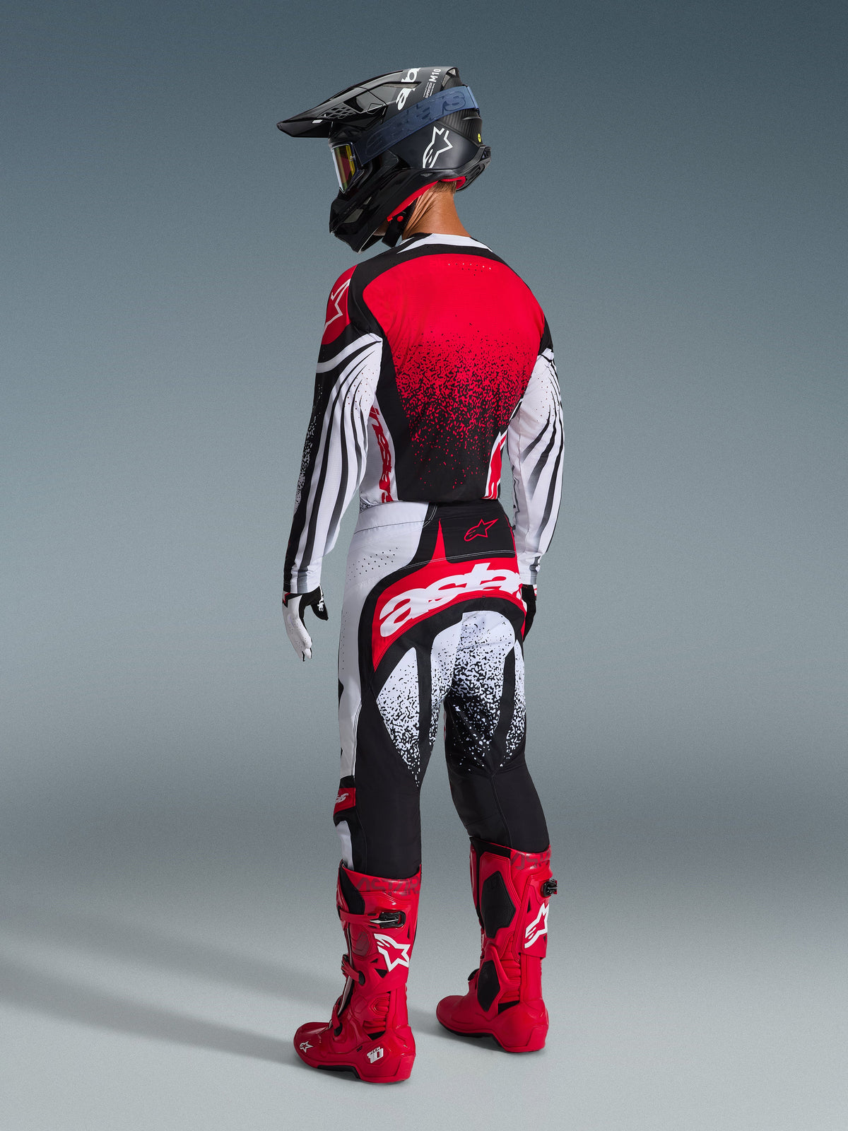 Zawodnik motocrossowy widziany od tyłu w spodniach Alpinestars 2026 Techstar Nomur w kolorze czarno-biało-czerwonym, duże białe logo „astars” na siedzeniu, dopasowana koszulka, jaskrawoczerwone buty i czarny kask, profesjonalny zestaw do wyścigów off-road